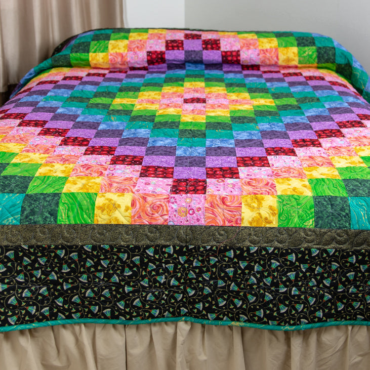A colorful quilted bedspread with a rainbow geometric pattern and a dark floral border, shown on a bed with a beige skirt in a bedroom.