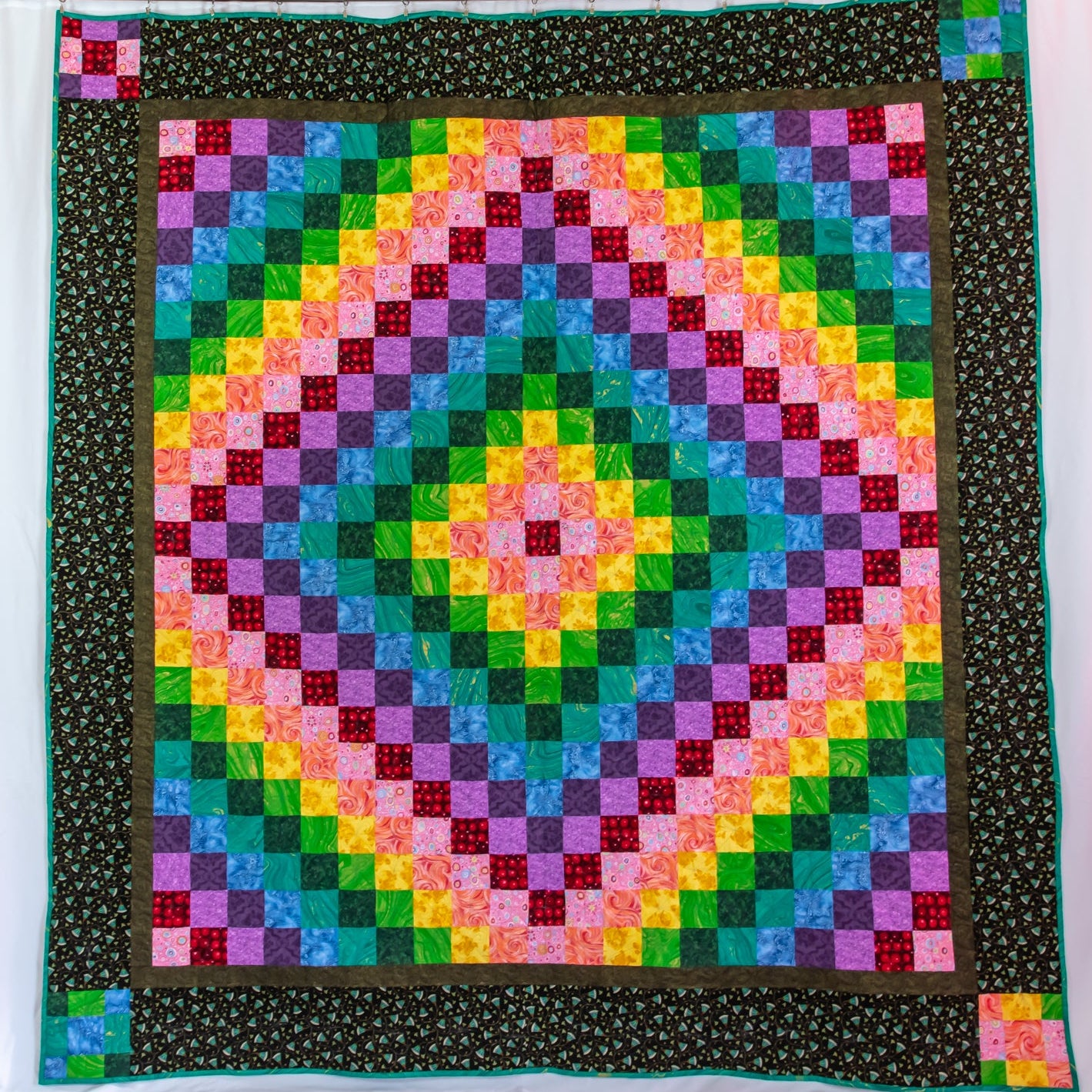 A square quilted textile with a colorful geometric medallion pattern in concentric rings of pink, yellow, green, blue, and purple, surrounded by a dark patterned border and a small coordinating patch on the lower right corner.