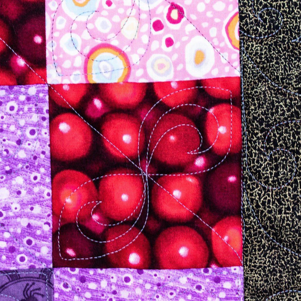 A close-up of a patchwork quilt or fabric panel with multiple square sections showing different patterns: red fruit-like dots, pink circles and rainbow motifs, purple dotted and floral designs, a black-and-white speckled section, and a narrow multicolor bird-patterned strip on the right.
