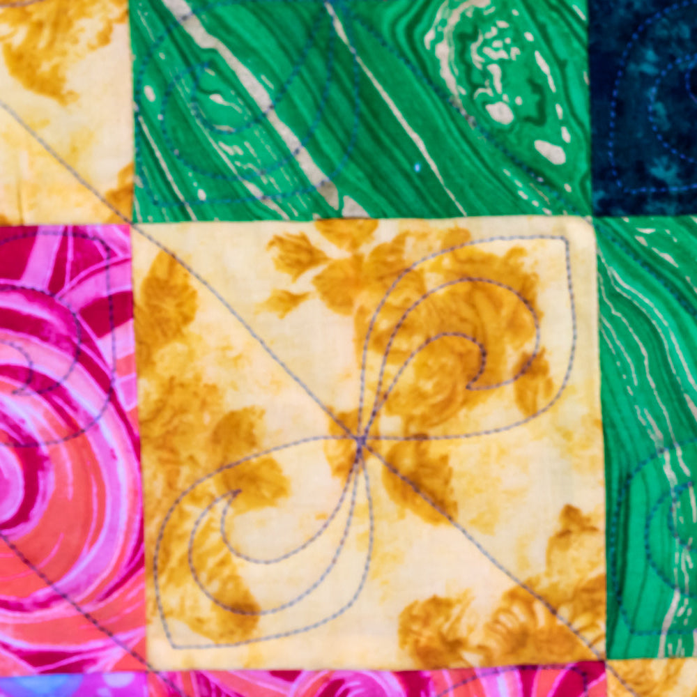A close-up of a colorful quilted fabric with a patchwork design. The fabric features various patterns including swirls, marbled veins, and floral prints in vibrant shades of green, blue, pink, yellow, and orange, with blue running stitch quilting visible on the center panels.
