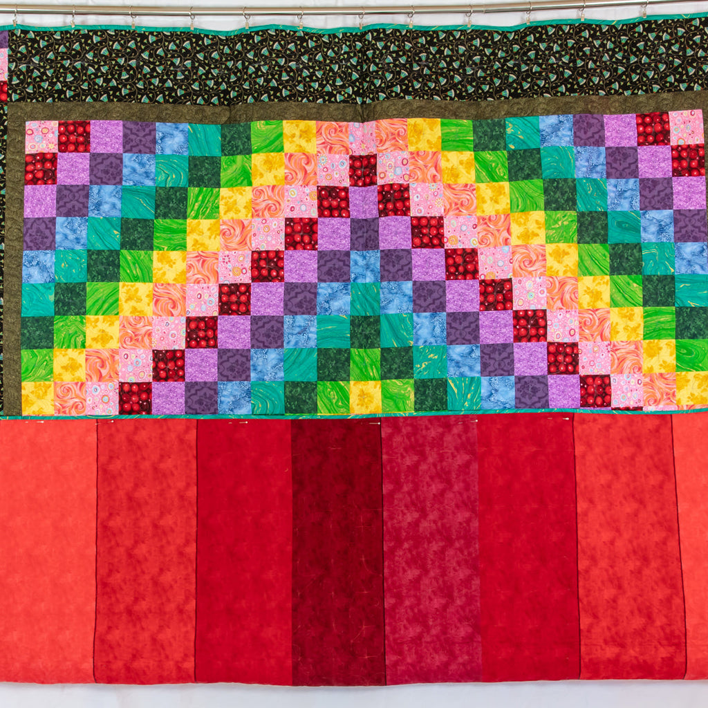 A handmade quilt featuring a rainbow-colored patchwork center, dark patterned side borders, and vertical red and coral stripes along the bottom, displayed against a white fabric backdrop.