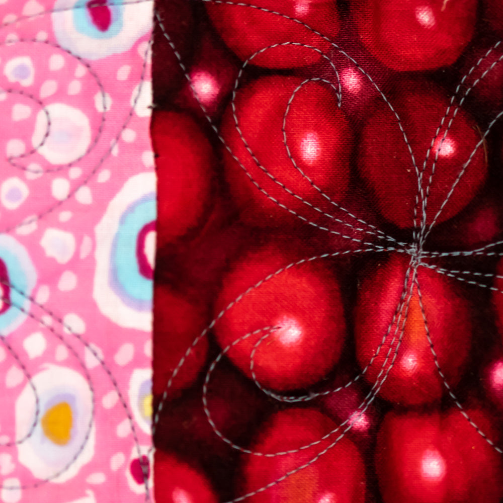 A close-up of a quilted fabric panel featuring a central design of glossy red cherries with swirling silver-gray stitched outlines, flanked by pink patterned borders with white, blue, and yellow motifs.