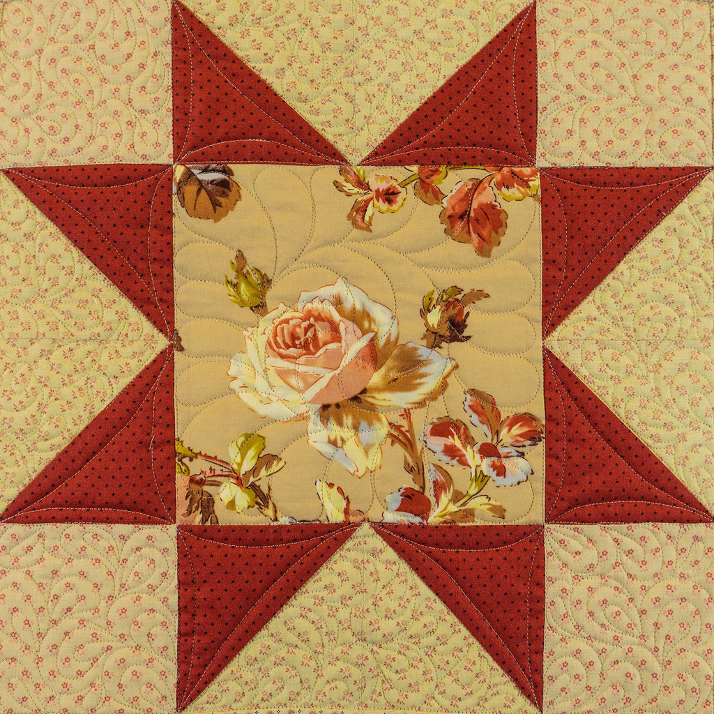 A square quilted patchwork star design featuring red star points and a central floral fabric panel with a large peach rose and smaller flowers on a beige background.