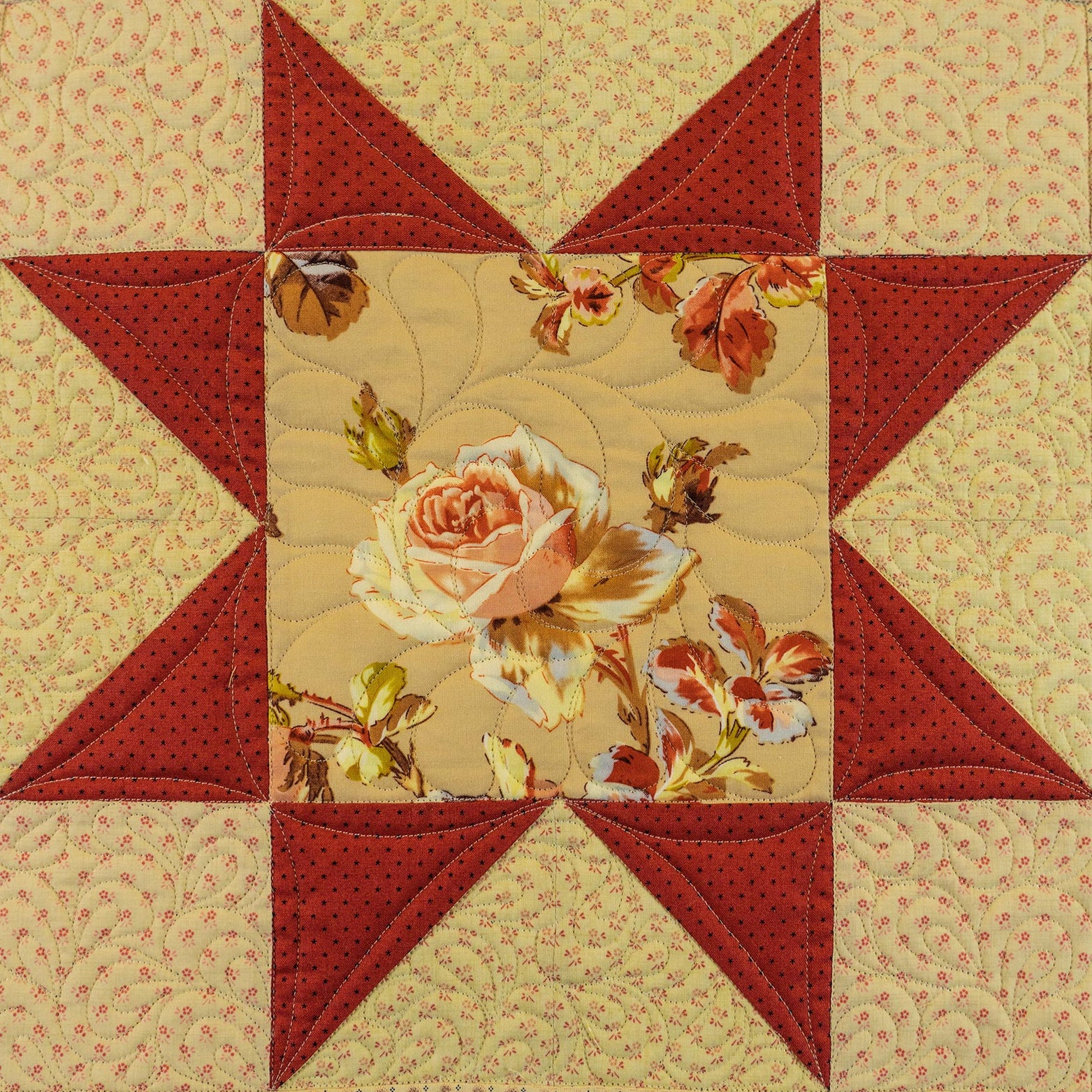 A square quilted patchwork star design featuring red star points and a central floral fabric panel with a large peach rose and smaller flowers on a beige background.