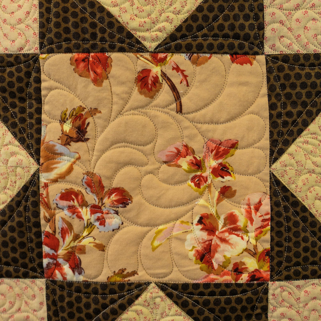 A close-up of a quilted fabric patchwork design featuring a central beige panel with a floral print in red, orange, and cream, surrounded by dark brown patterned triangles and cream swirl-patterned sections.