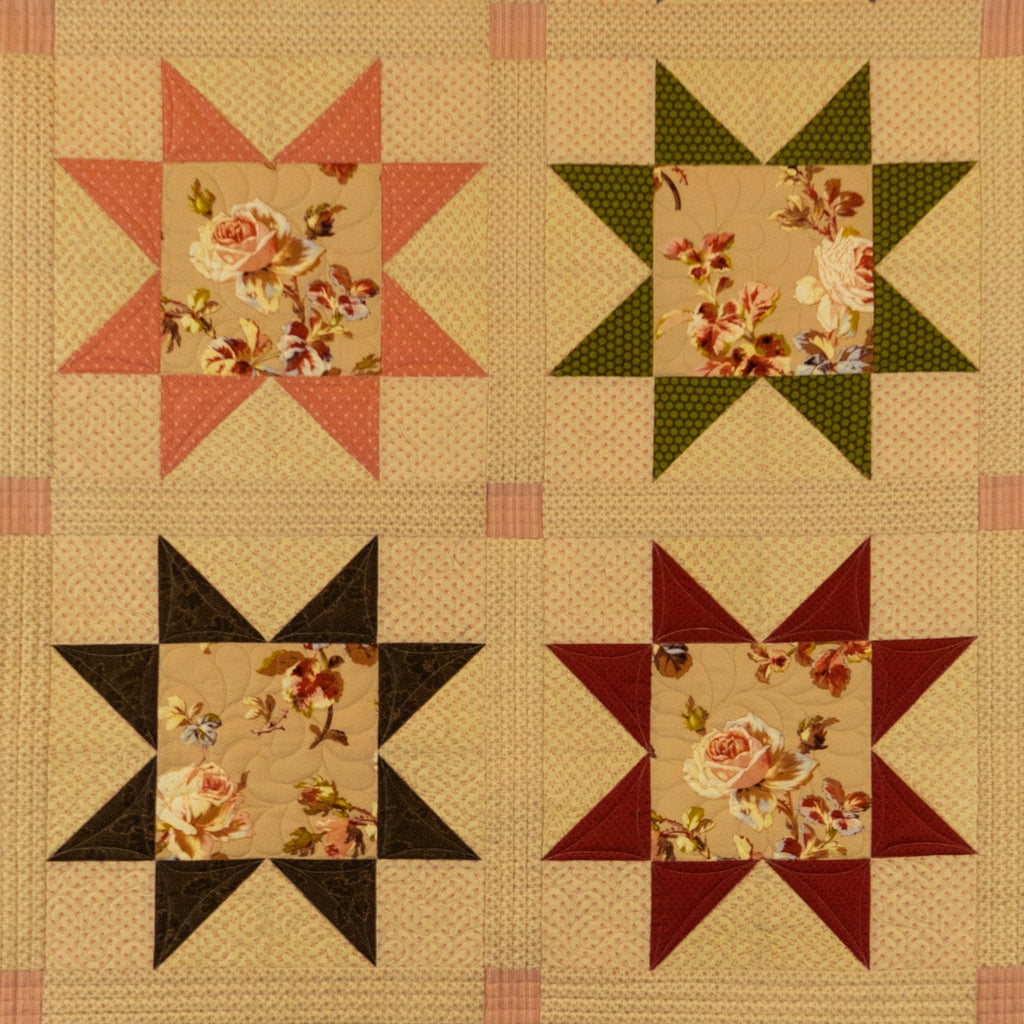 A vintage-style quilt or fabric panel featuring four starburst blocks arranged in a 2x2 grid. Each block has a floral center with surrounding solid sections in pink, green, black, and burgundy on a beige background.