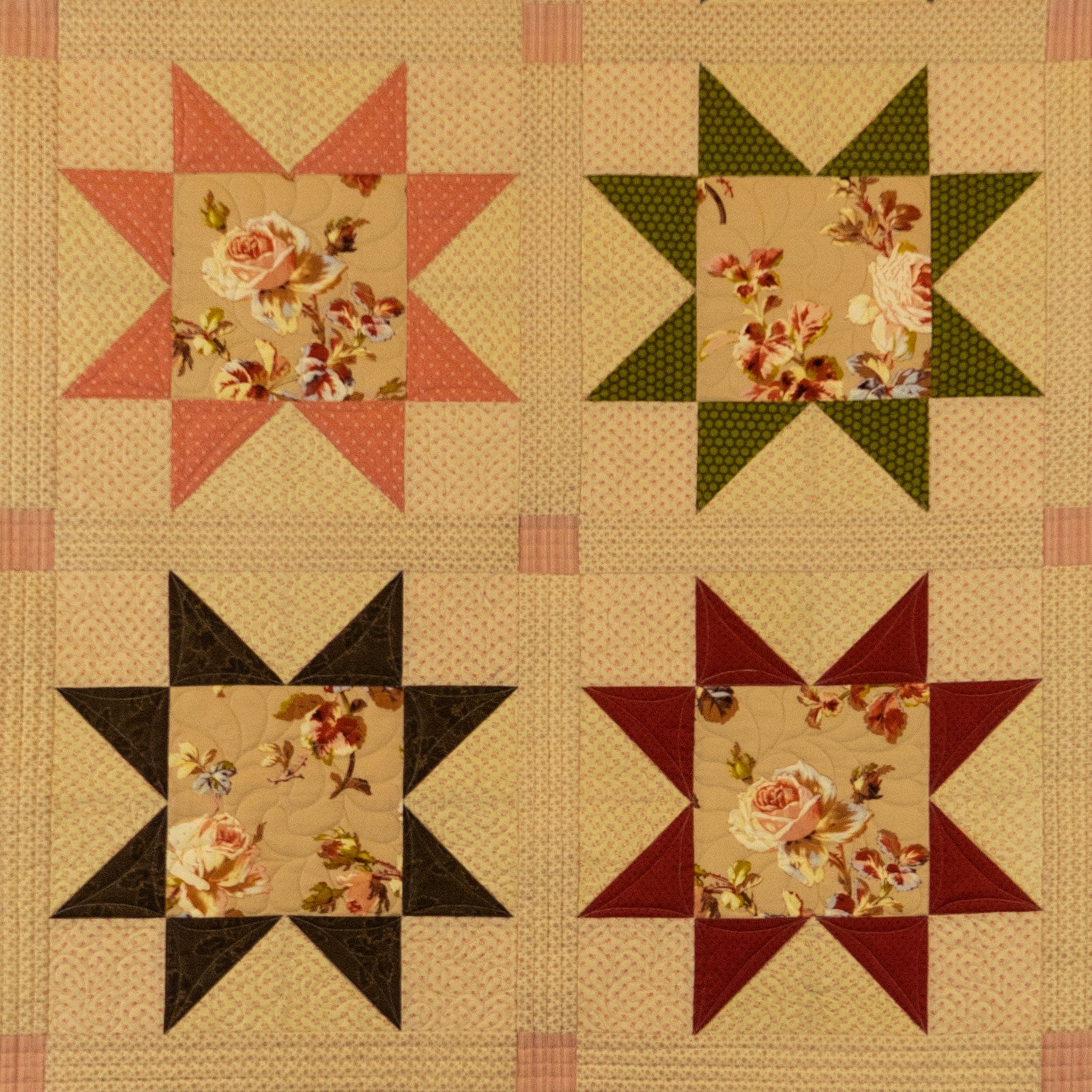 A vintage-style quilt or fabric panel featuring four starburst blocks arranged in a 2x2 grid. Each block has a floral center with surrounding solid sections in pink, green, black, and burgundy on a beige background.