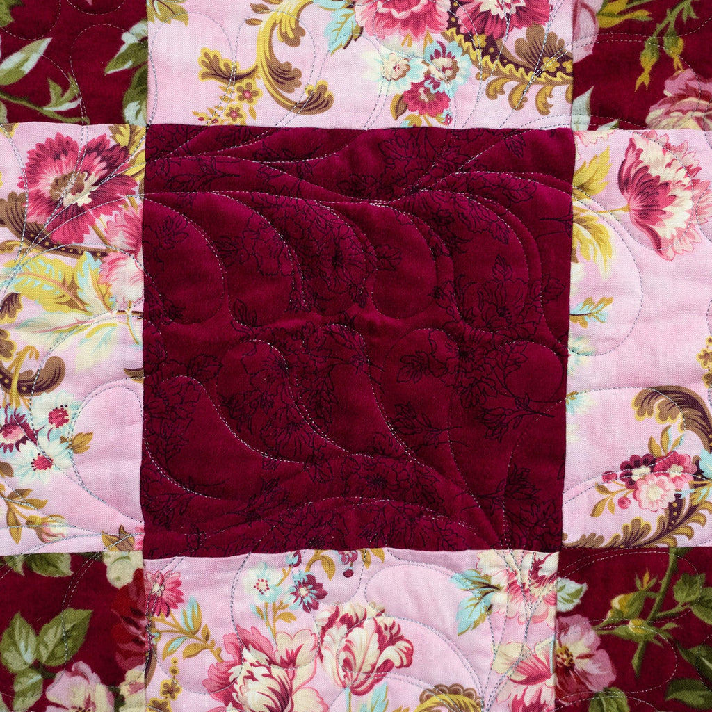 A close-up of a quilted textile featuring a central square of deep burgundy velvet-like fabric surrounded by floral patchwork panels in pink, burgundy, green, and cream.