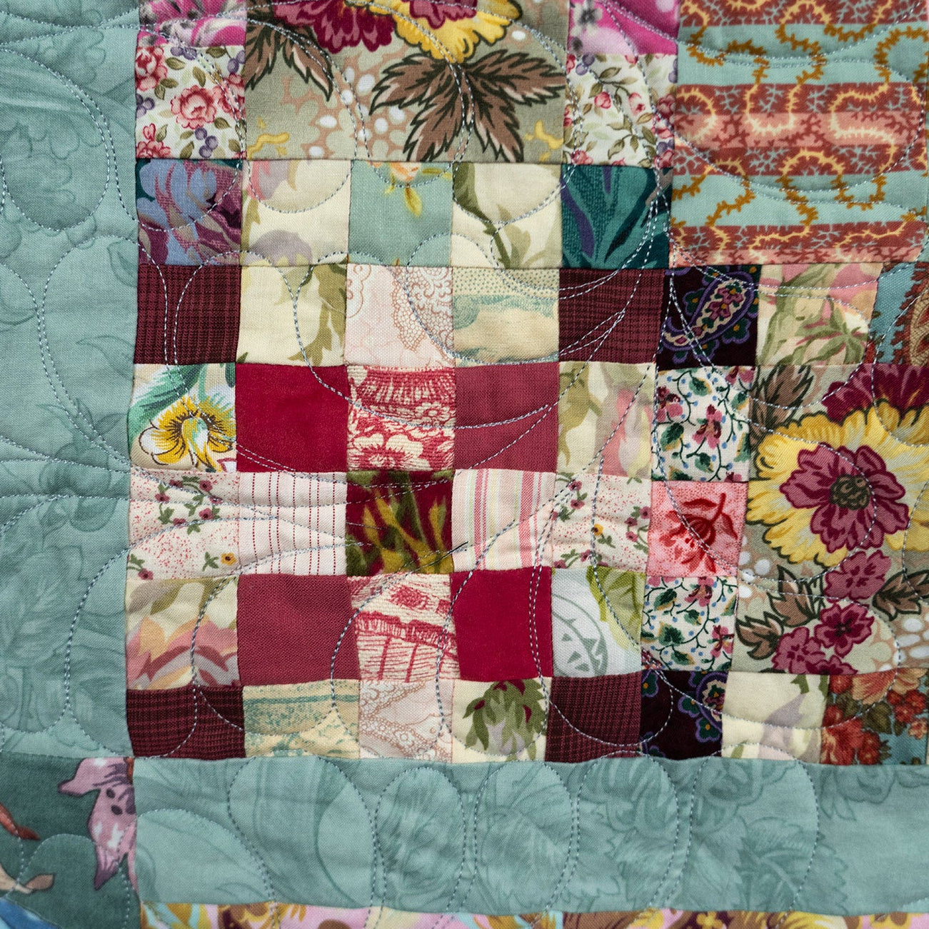 A close-up of a patchwork quilt featuring a central grid of floral and patterned fabric squares in pink, red, green, cream, and yellow, bordered by solid aqua blue fabric.