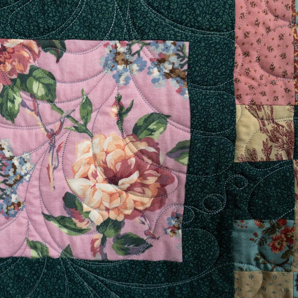 A close-up of a quilted textile featuring a central pink floral patch with large peach and cream roses, green leaves, and small blue flowers, surrounded by a dark teal quilted border and a patchwork side panel.