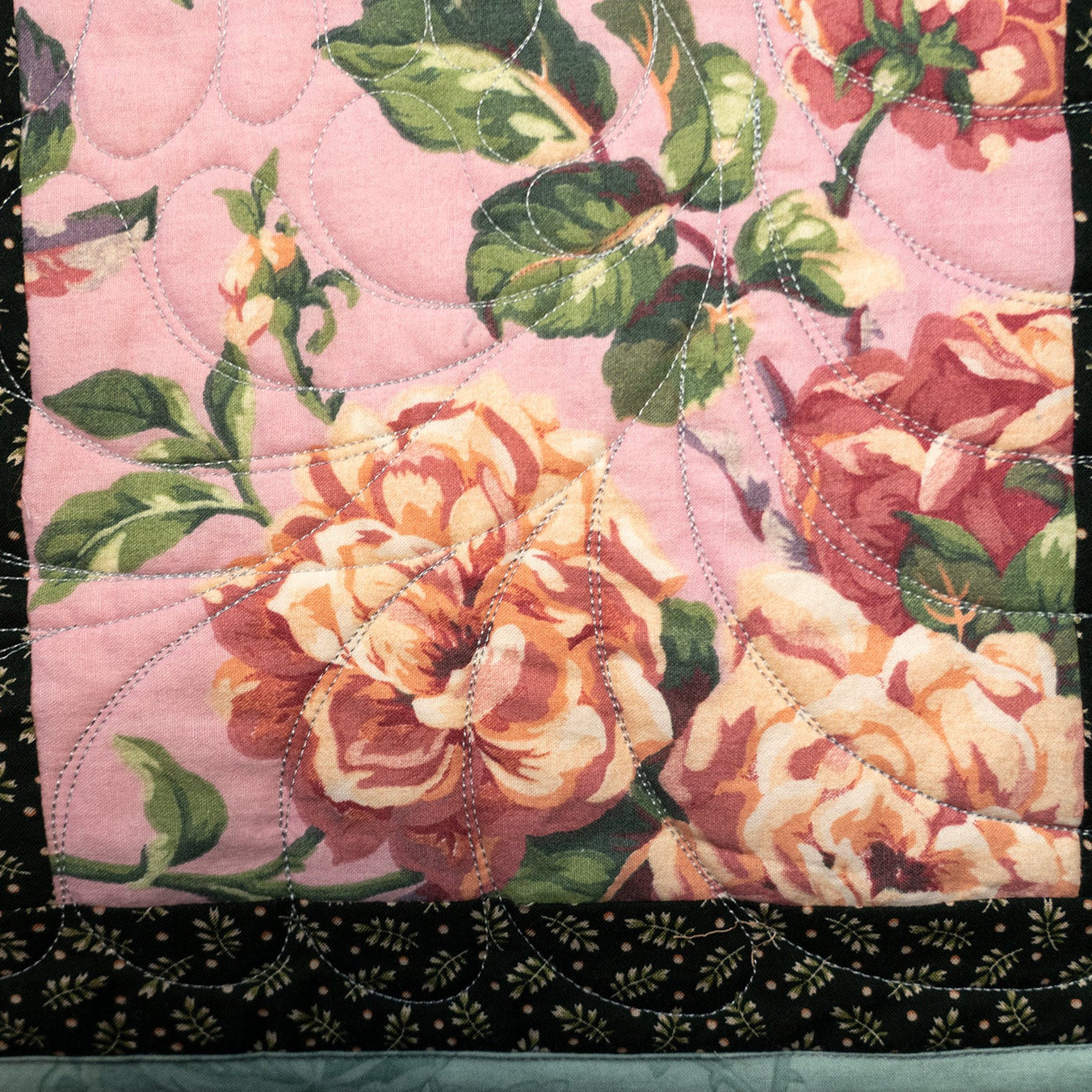 A close-up of a quilted textile featuring a pink floral panel with large peach and cream roses, green leaves, and a black border with small gold floral motifs and a light blue edge.