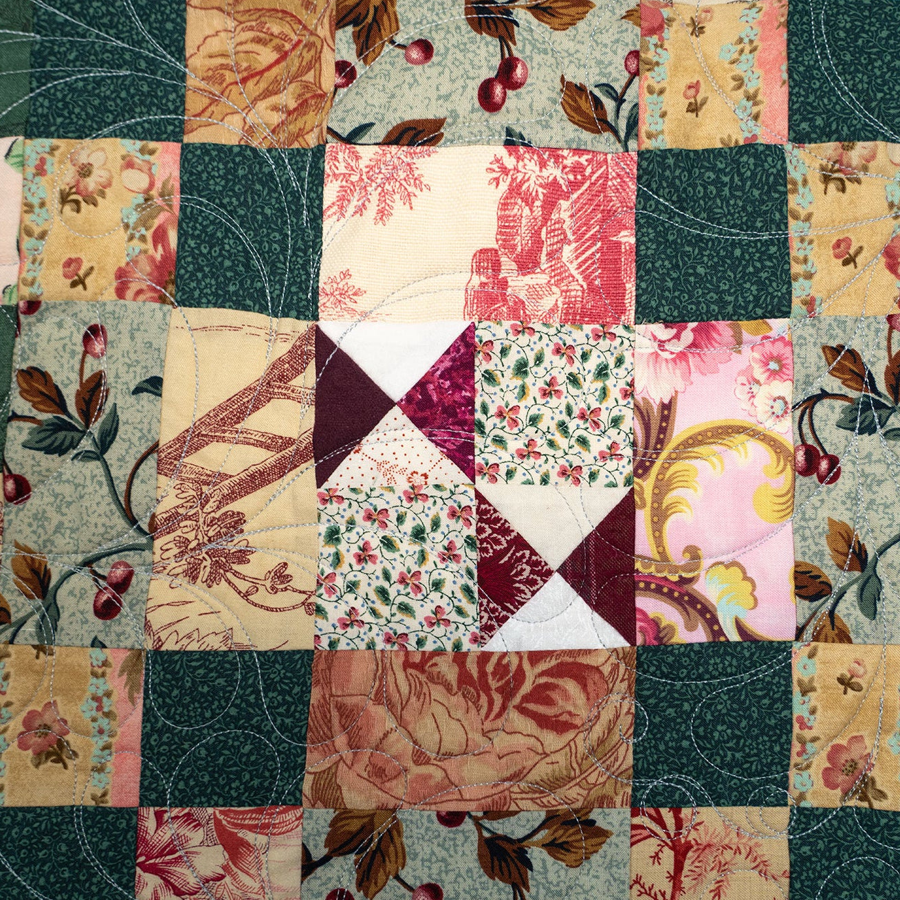 A square patchwork quilt featuring a central diamond-shaped block with alternating dark red and cream triangles, surrounded by floral and botanical patterned fabric squares in pink, green, beige, and teal.