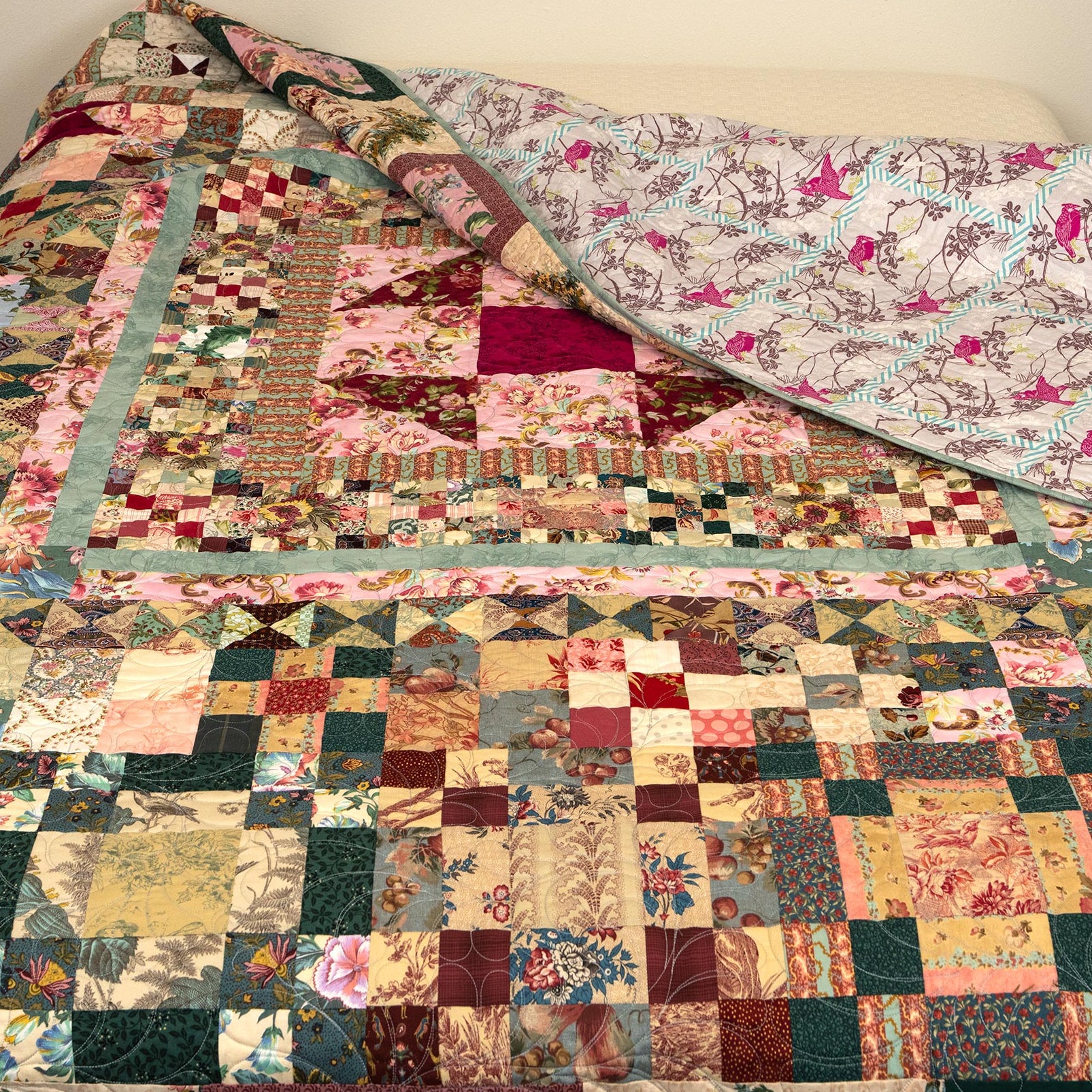 A handmade patchwork quilt draped over a bed, showing a complex multi-colored design with floral and geometric blocks, with the back side featuring a light blue and pink bird pattern.