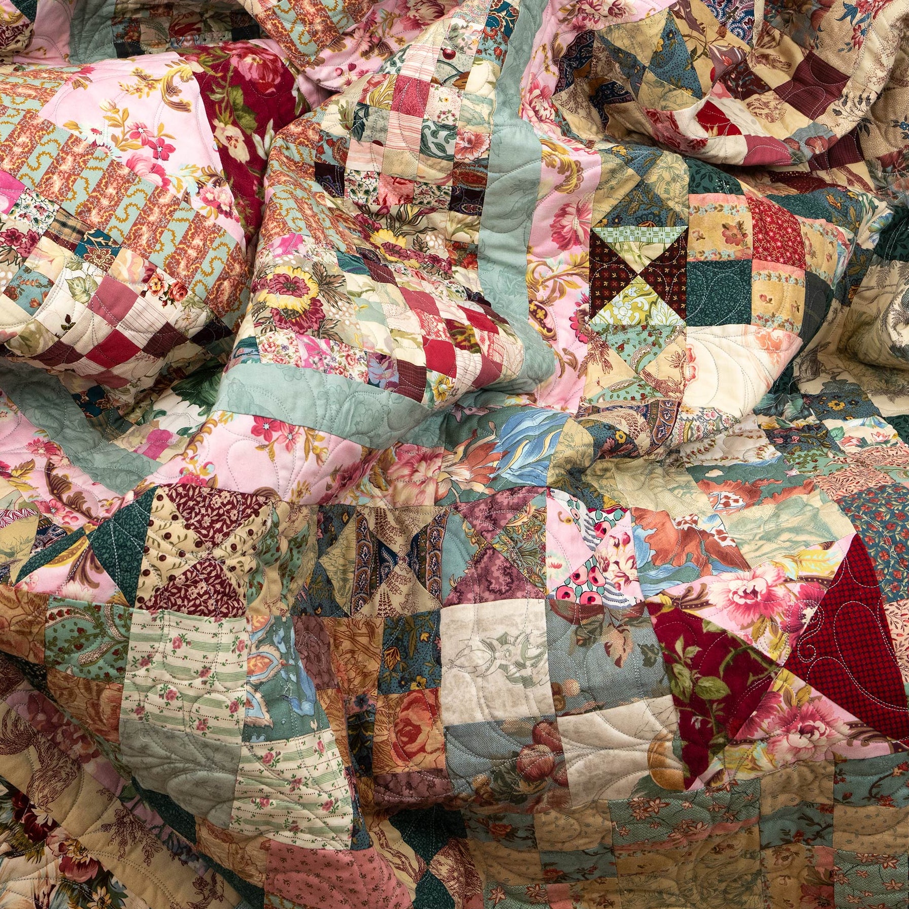 A close-up of a patchwork quilt with multiple fabric panels in pink, teal, red, beige, and floral prints, filling the frame.