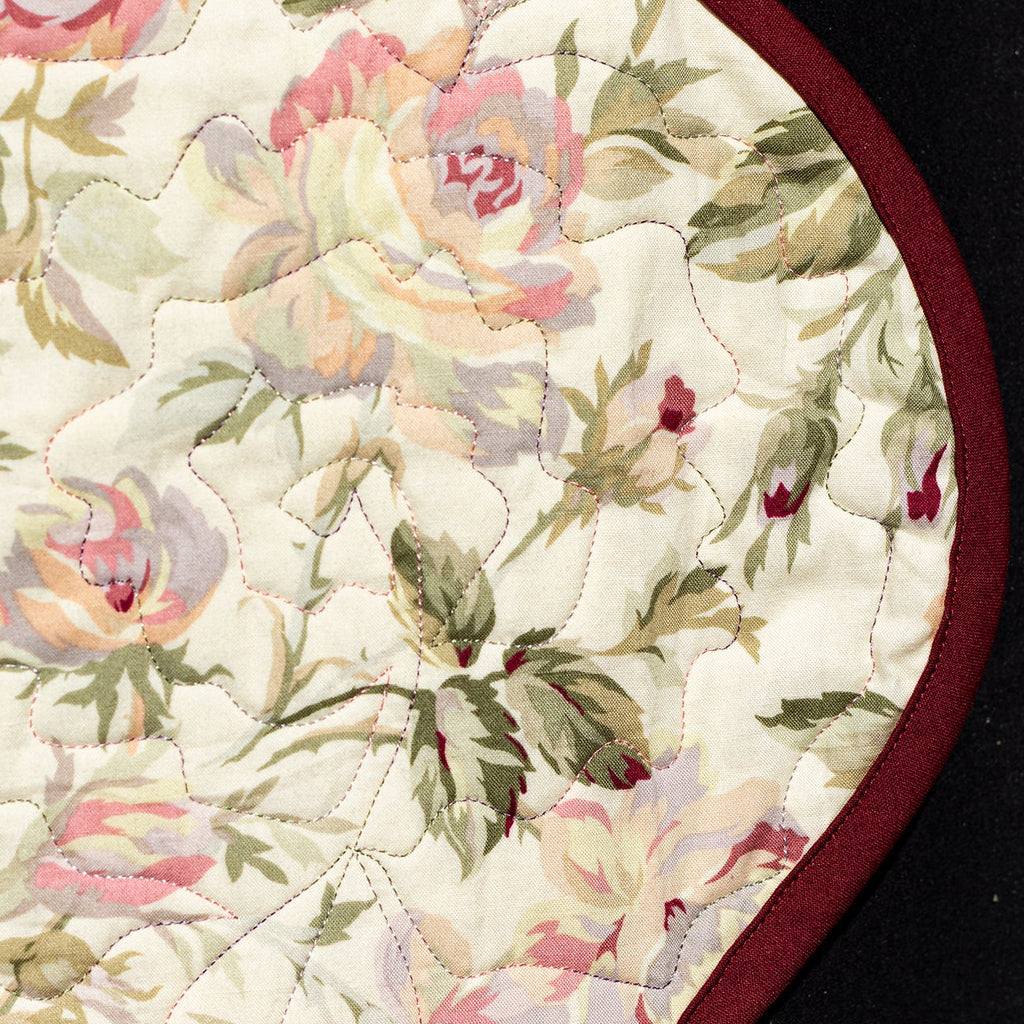 A close-up of a vintage floral pattern with pink, peach, and green roses on an off-white background, finished with a dark red stitched border, shown against a black background.
