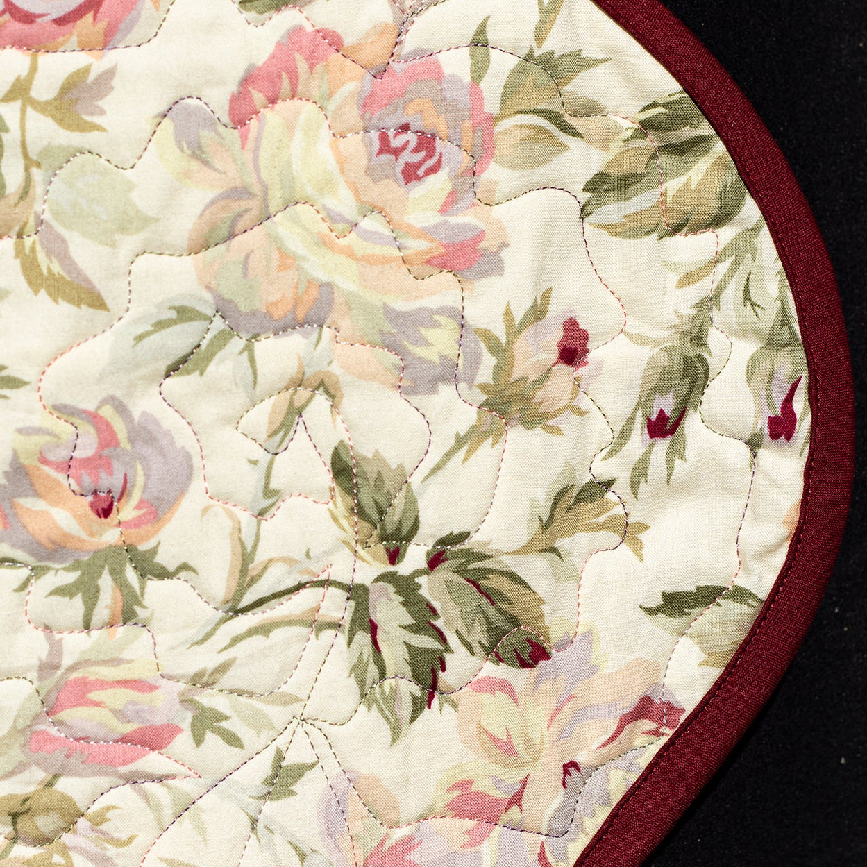 A close-up of a vintage floral pattern with pink, peach, and green roses on an off-white background, finished with a dark red stitched border, shown against a black background.