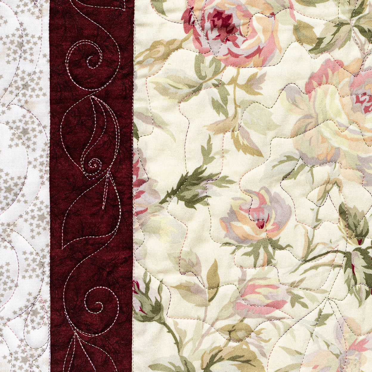 A close-up of a quilted textile featuring a cream floral print with pink and green roses, a dark red decorative border with white swirl stitching, and a white patterned section on the left.
