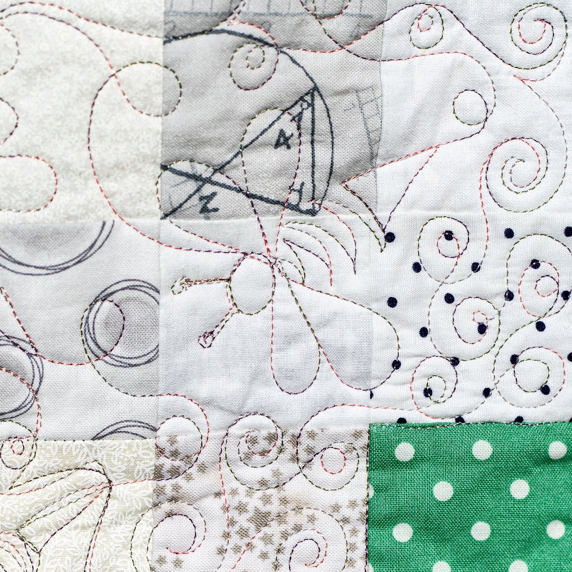 A close-up of a quilted fabric panel with mixed patchwork sections, including white and gray swirl quilting, a green square with white polka dots, and a central gray printed patch. The image is bordered by black space on the left and right.