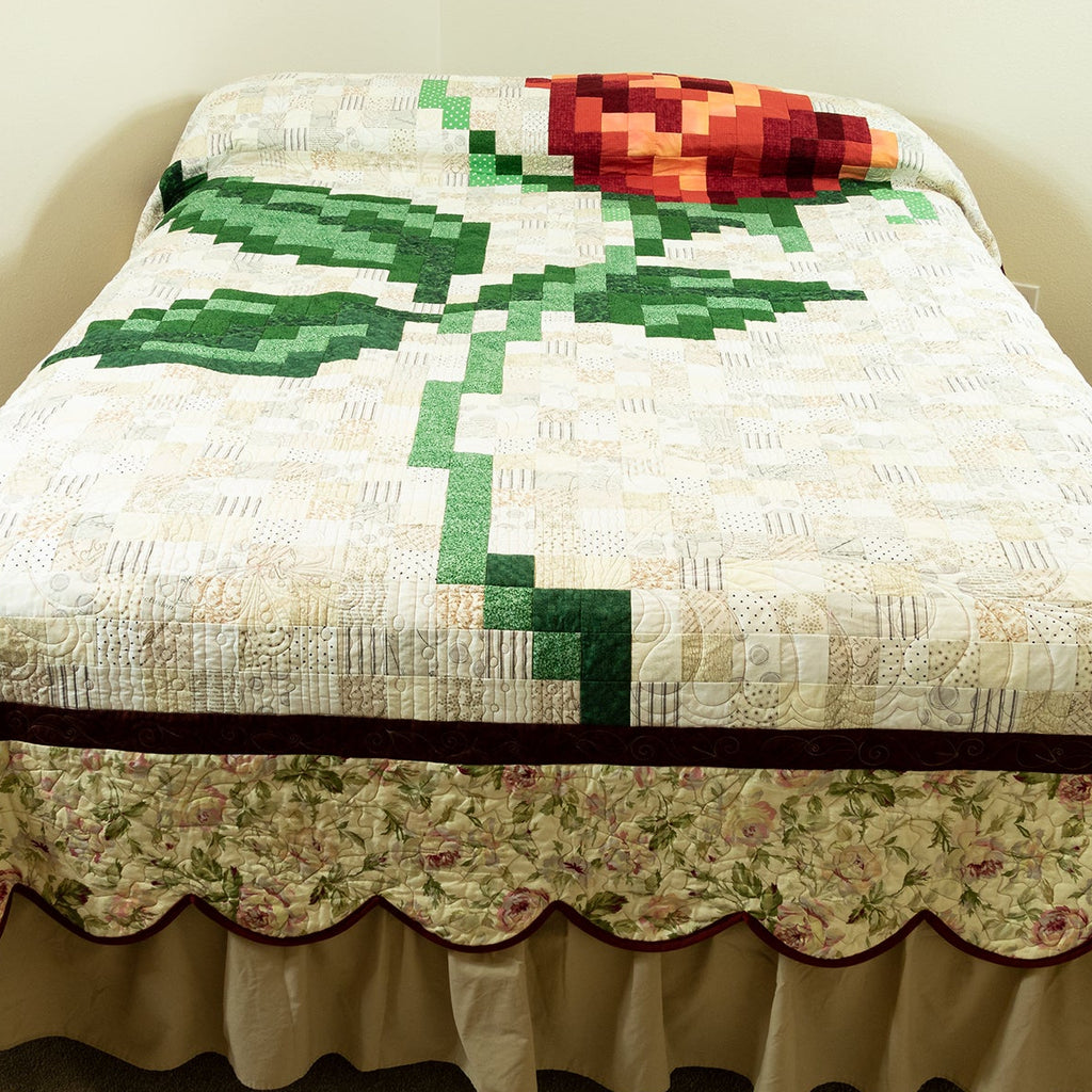 A quilted bedspread featuring a large pixelated green rose motif on a cream background, with a floral scalloped border and beige ruffled skirt, shown on a bed in a simple room.