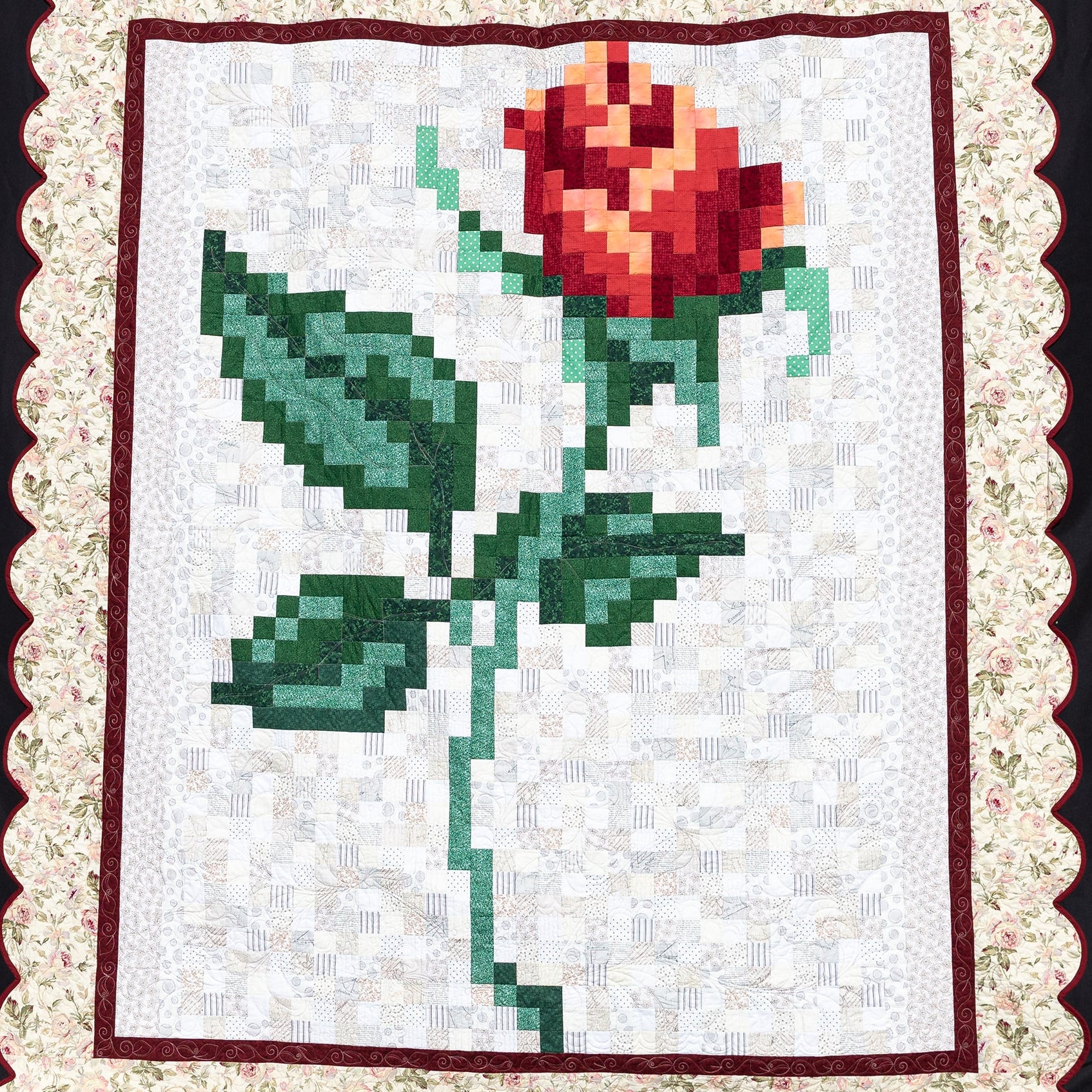 A square quilted patchwork artwork featuring a pixelated-style red rose with green leaves on a white background, surrounded by a scalloped floral border and dark red inner trim, shown against a black backdrop.