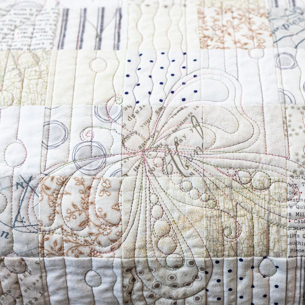 A close-up of a patchwork quilt featuring various fabric panels with quilting stitches, including script, stripes, dots, and floral motifs in soft beige, cream, and gray tones.