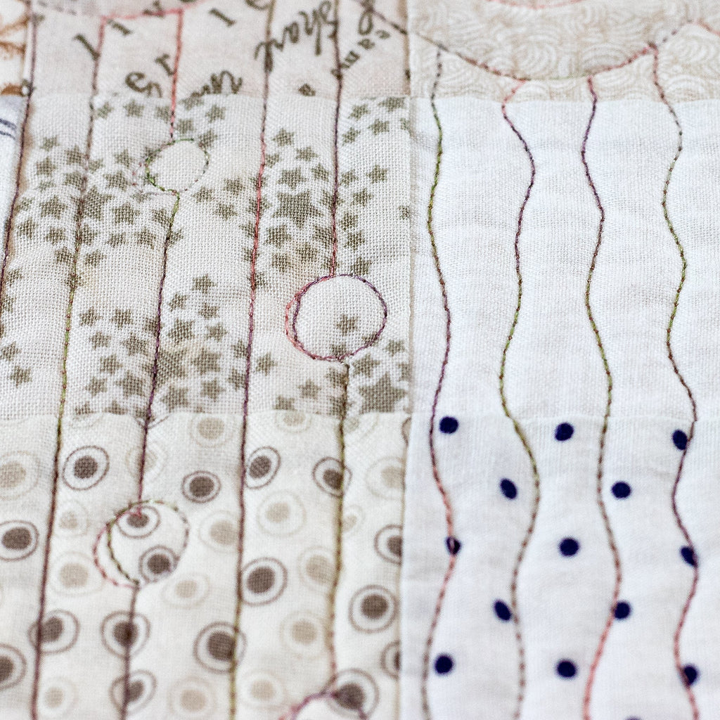 A close-up of a handmade quilted fabric panel featuring various stitched patterns including vertical wavy lines with blue dots, vertical lines with star prints, and circular dot motifs on a white and beige background.