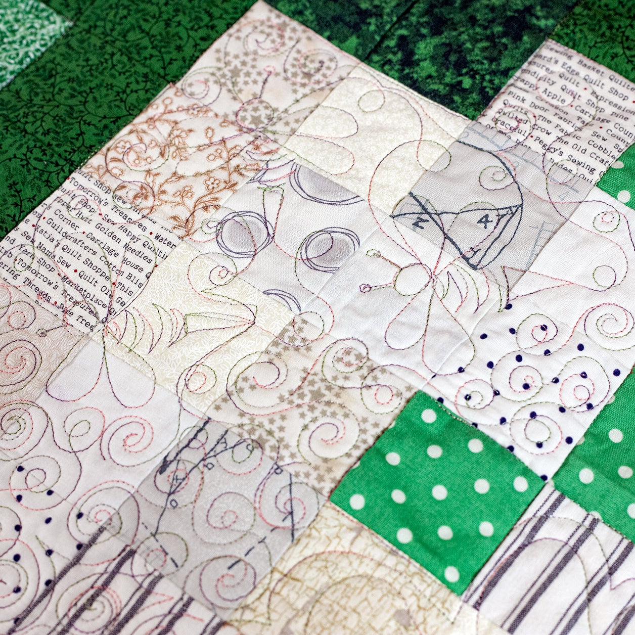 A close-up of a patchwork quilt featuring various fabric patterns, including green polka dot, green textured blocks, and cream sections with printed text and quilting designs.