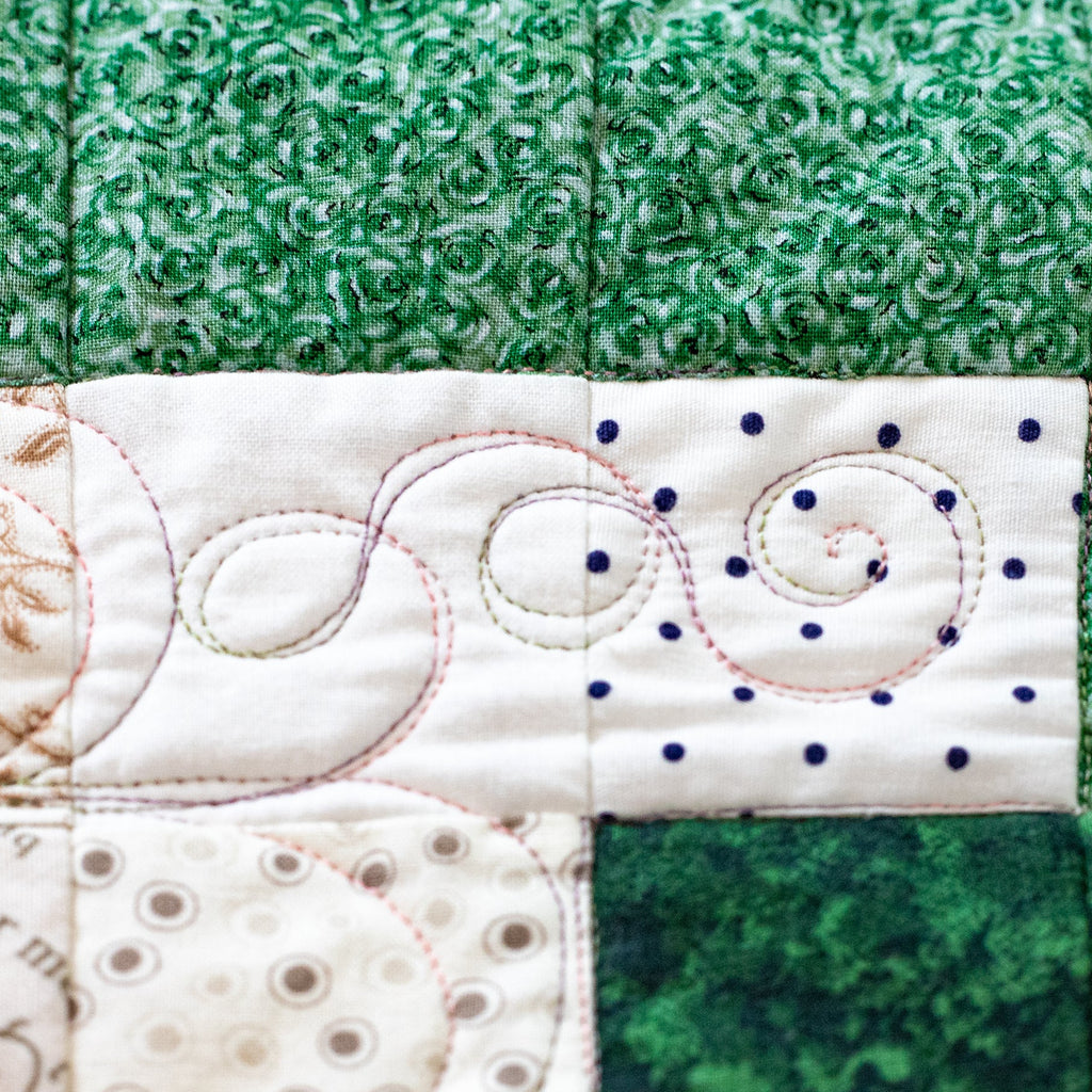 A close-up of a quilted fabric panel featuring green and cream patterned sections with decorative stitching, including swirls and dotted lines.