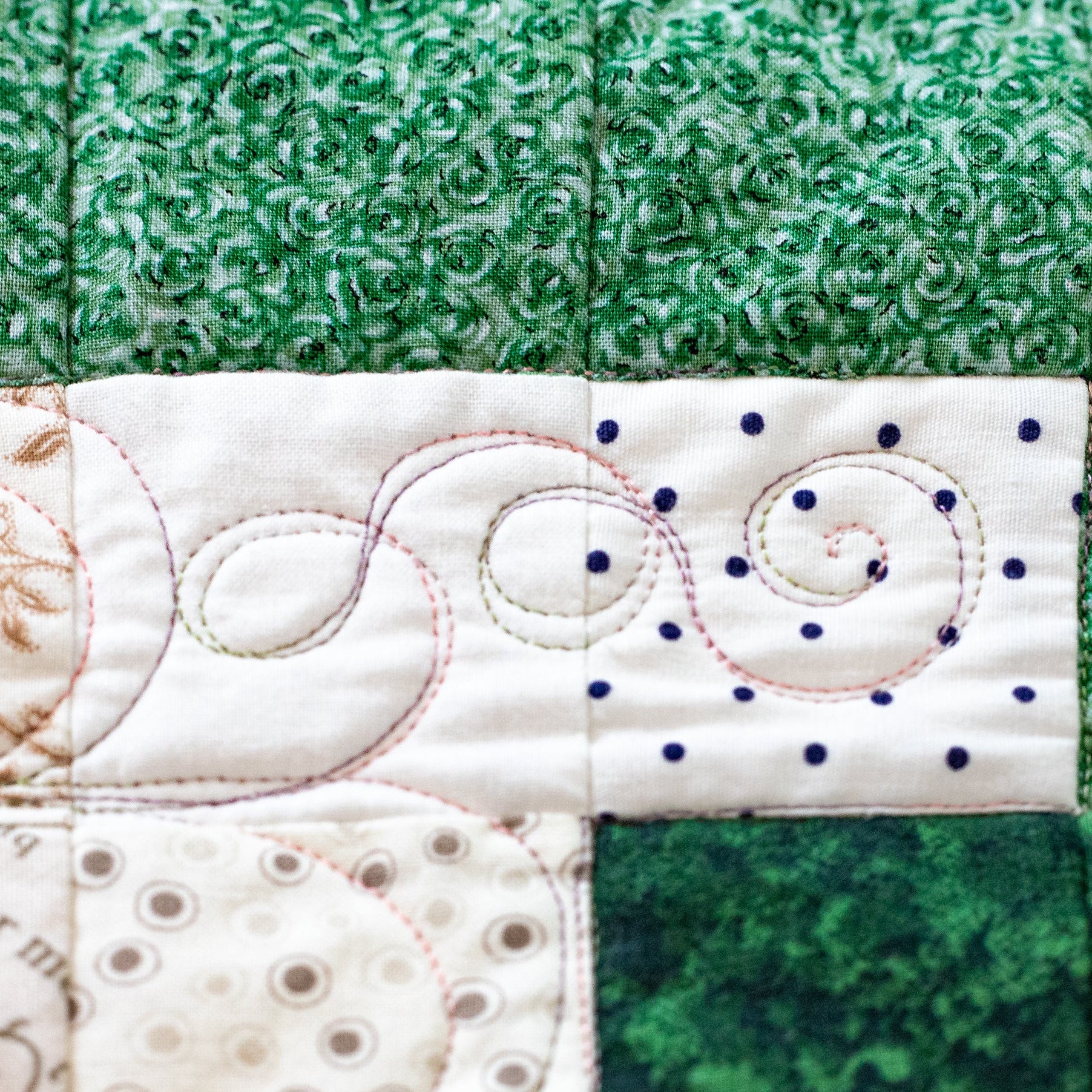 A close-up of a quilted fabric panel featuring green and cream patterned sections with decorative stitching, including swirls and dotted lines.
