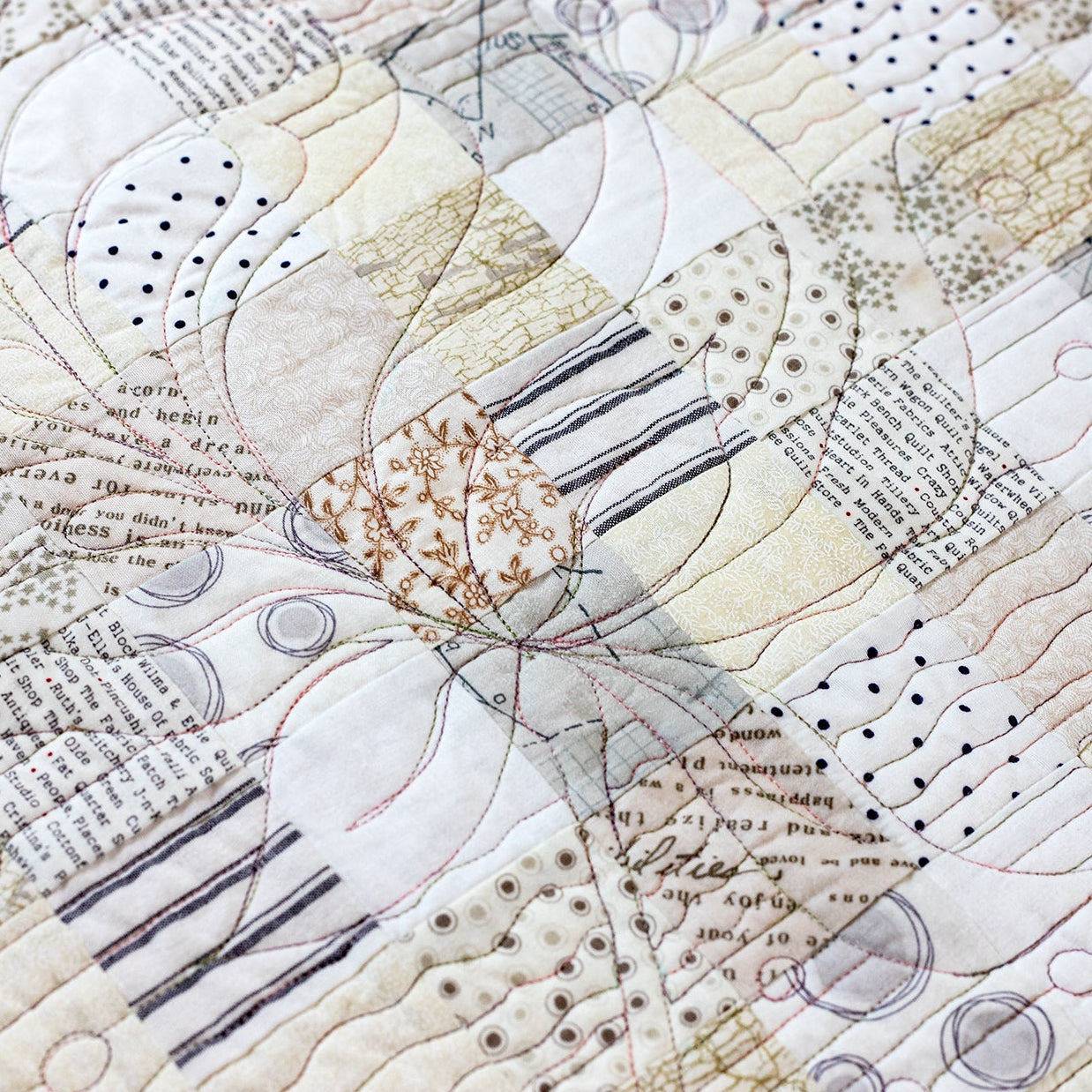 A close-up of a quilted fabric featuring a patchwork design with floral and abstract motifs in beige, cream, brown, and gray tones, with stitched text details.