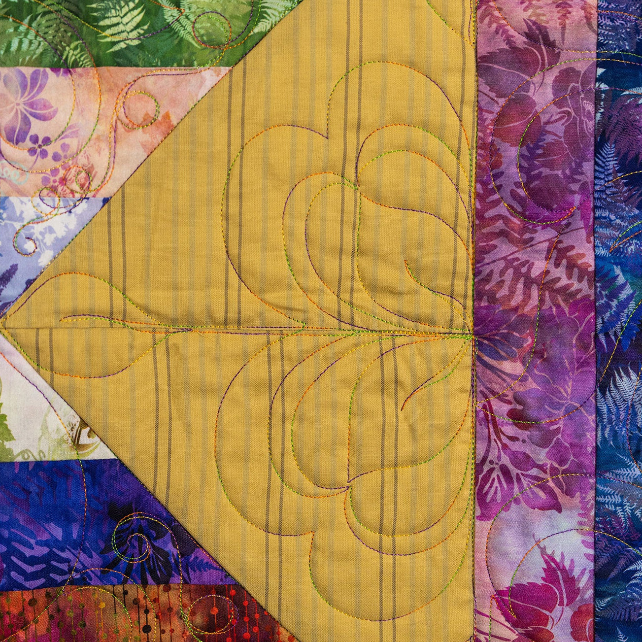A close-up of a colorful quilted textile with geometric and floral fabric panels, including a large yellow section with stitched leaf quilting, surrounded by purple, blue, pink, green, and orange patterned fabrics.