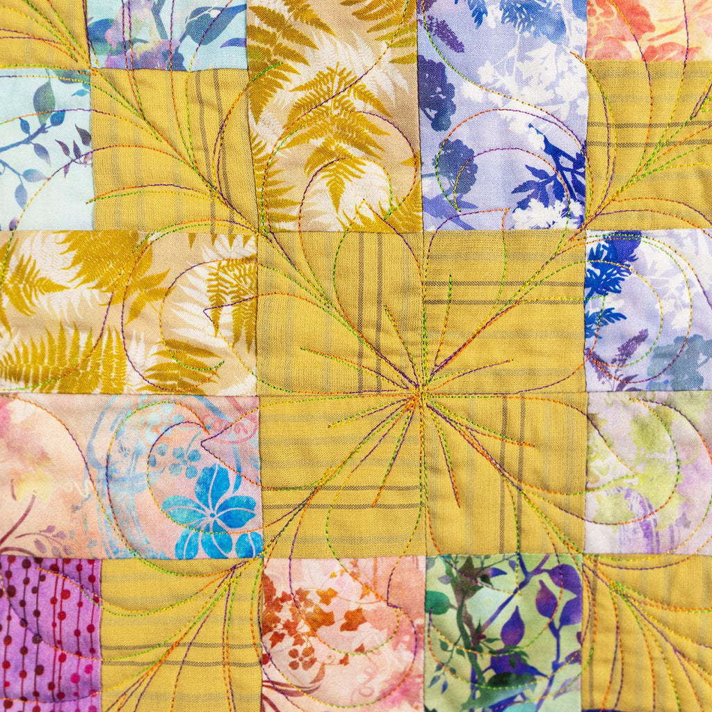 A square patchwork quilt featuring a central yellow starburst block surrounded by floral and botanical patterned fabric squares in yellow, purple, pink, blue, and green.
