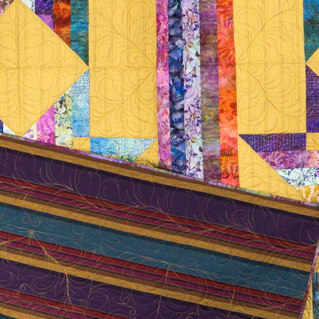 A close-up of a colorful quilted fabric surface with geometric patchwork panels in yellow, purple, blue, orange, and floral prints, plus decorative quilting lines and a striped border.