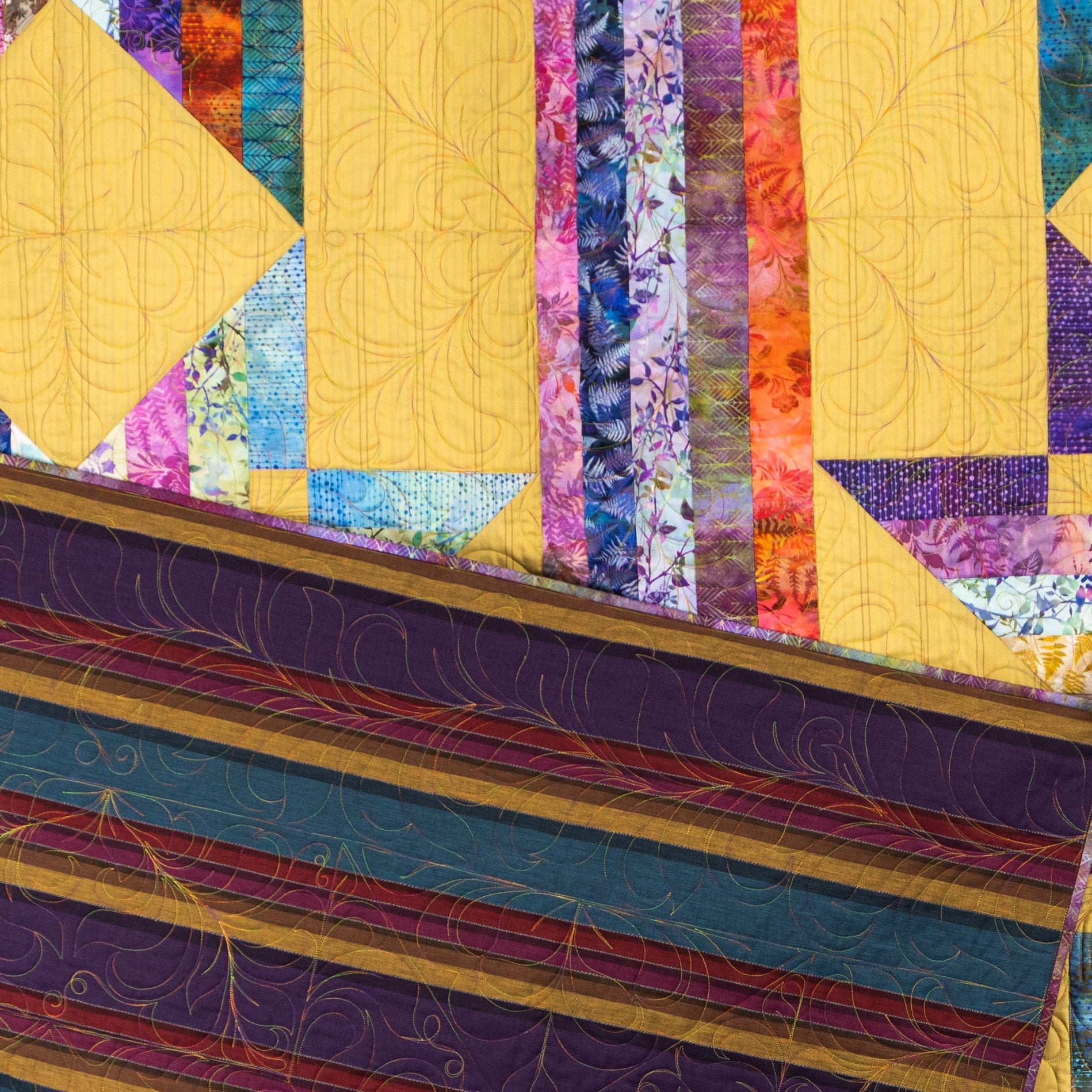 A close-up of a colorful quilted fabric surface with geometric patchwork panels in yellow, purple, blue, orange, and floral prints, plus decorative quilting lines and a striped border.