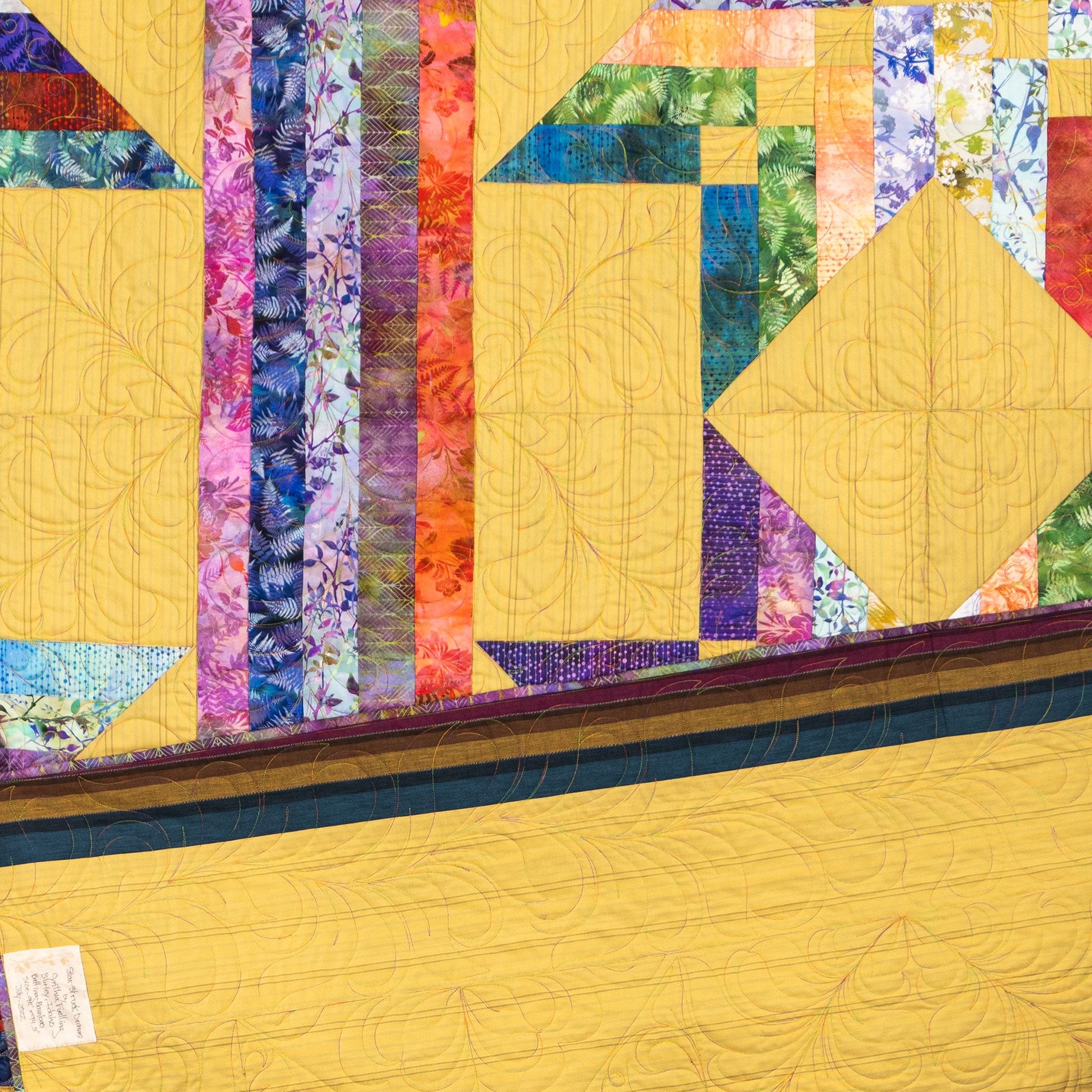 A close-up of a quilted fabric panel with a geometric patchwork design. The design features a large yellow field with stitched swirl patterns, vertical bands of multicolored floral and patterned fabric in purple, blue, green, orange, and pink, and a dark teal border.