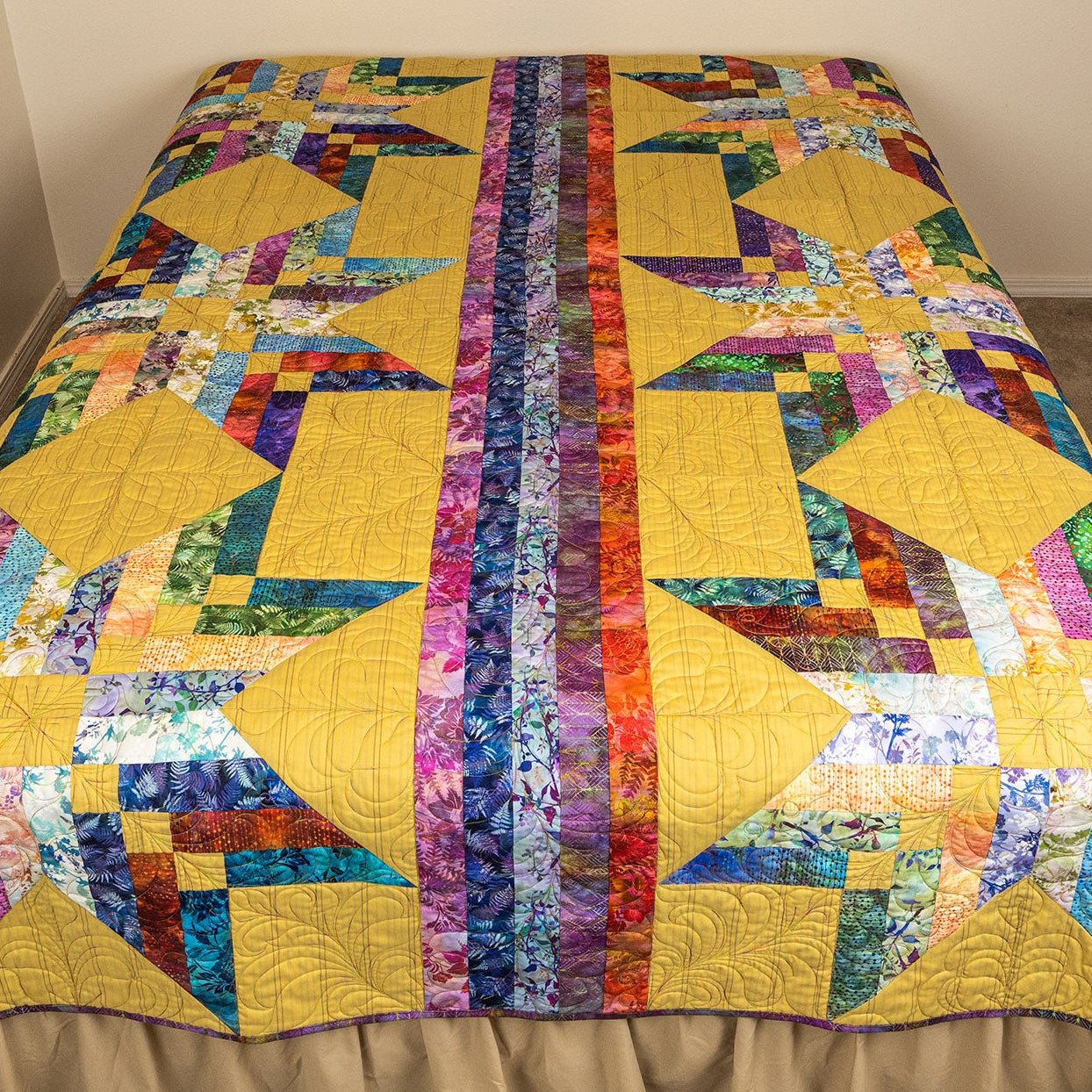 A colorful patchwork quilt with geometric star and block patterns, draped over a bed with a beige ruffled bed skirt in a bedroom setting.