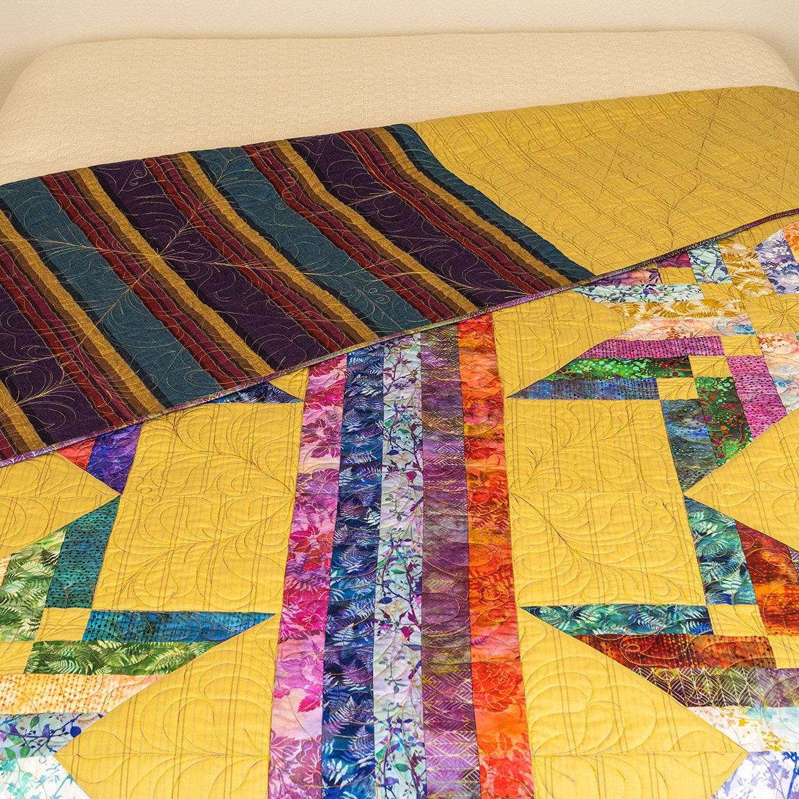A colorful patchwork quilt draped over a bed, featuring large yellow sections, striped bands, and floral patterned accents in blue, purple, green, orange, and pink.
