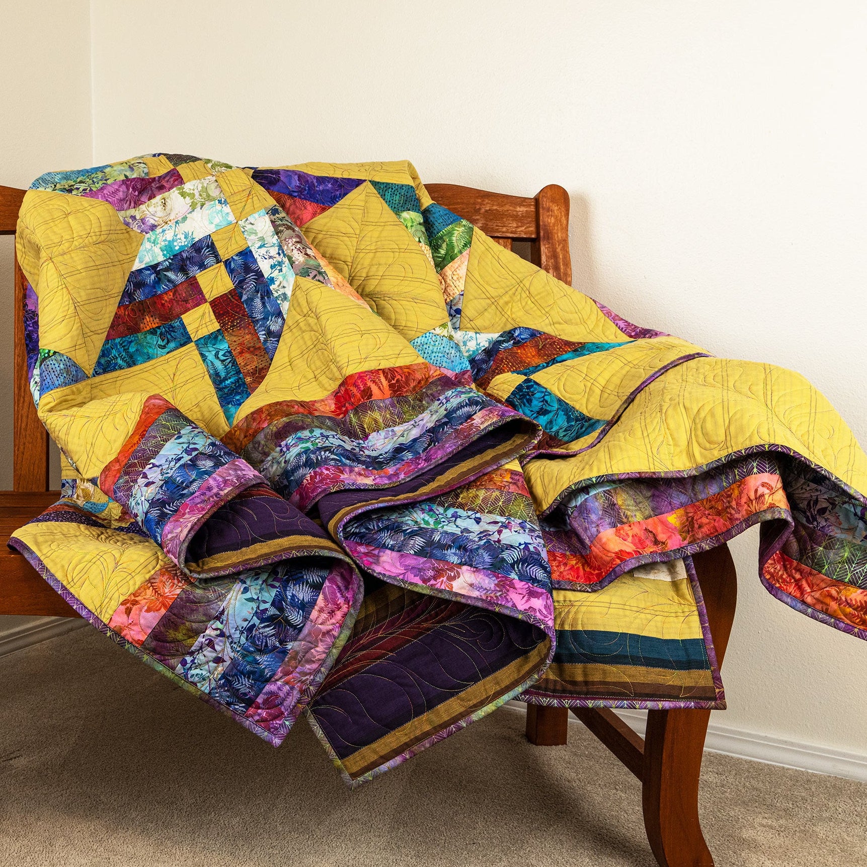 A colorful patchwork quilt with prominent yellow sections and blue, purple, and red patterns, draped over a wooden chair in a home setting.