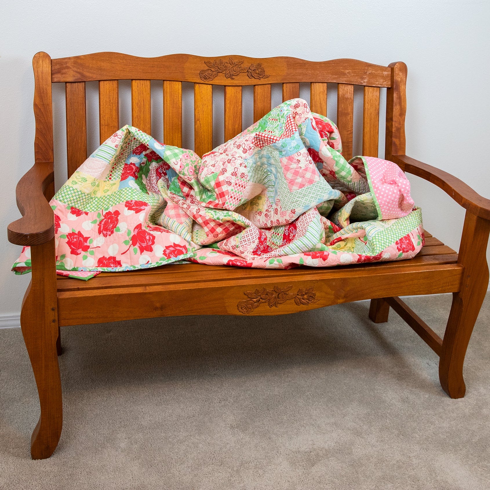Wooden bench with a floral quilt draped over it against a plain wall.