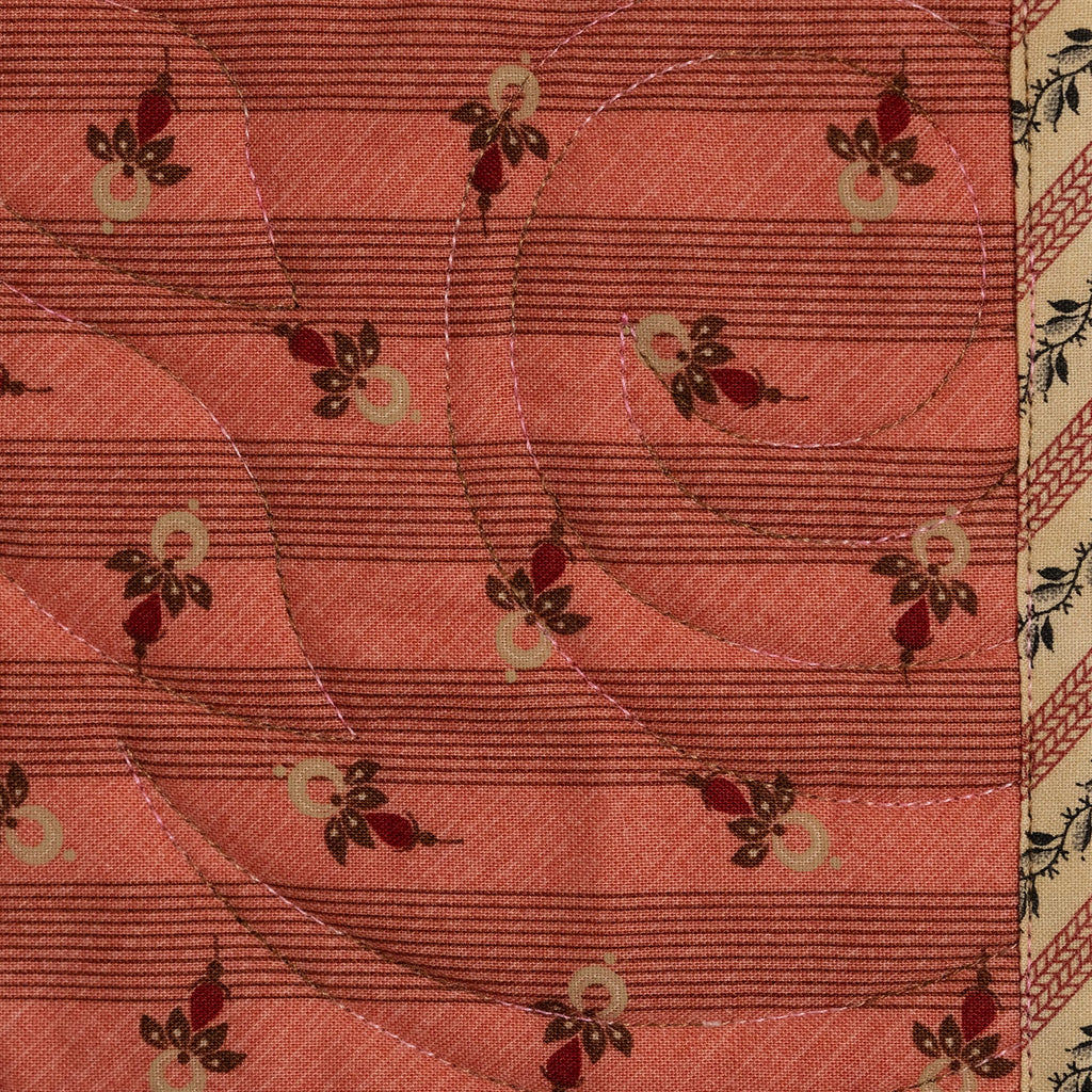 A close-up of a quilted fabric featuring a coral pink background with horizontal stitching, curved quilting lines, and small floral motifs in red, green, and cream. The fabric is bordered on the sides with patterned textile borders.