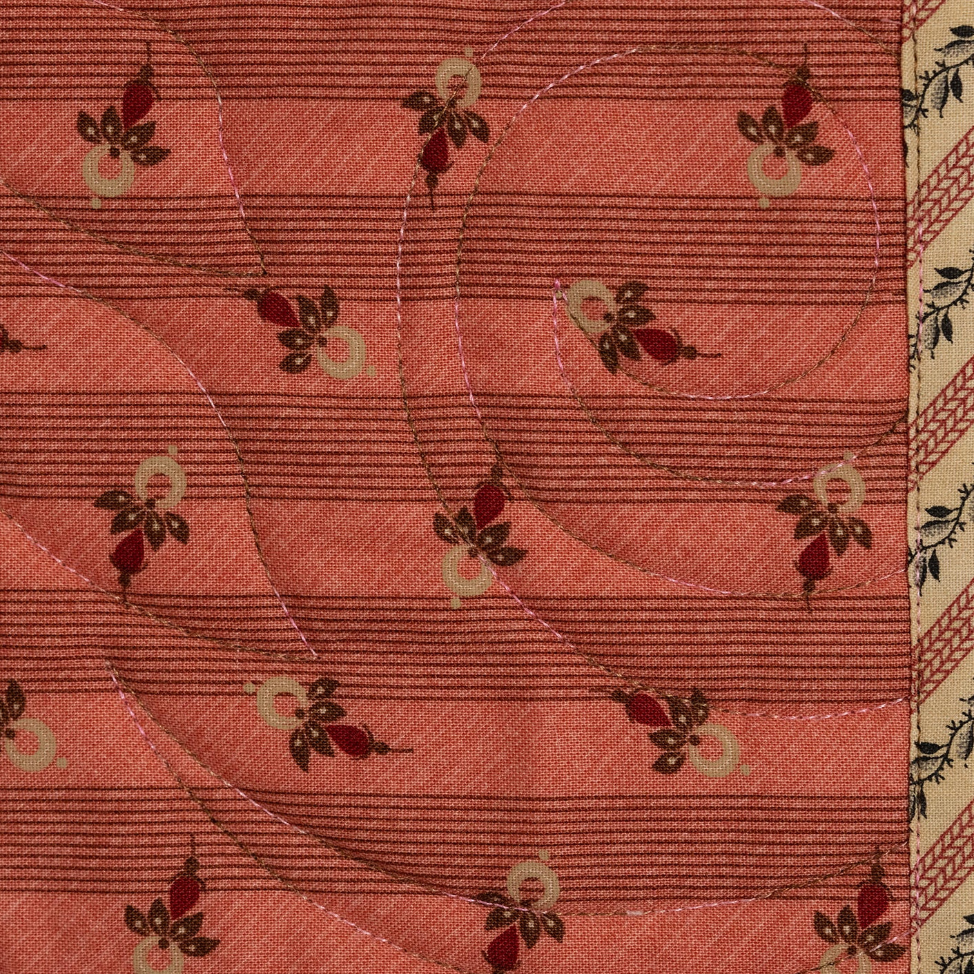 A close-up of a quilted fabric featuring a coral pink background with horizontal stitching, curved quilting lines, and small floral motifs in red, green, and cream. The fabric is bordered on the sides with patterned textile borders.
