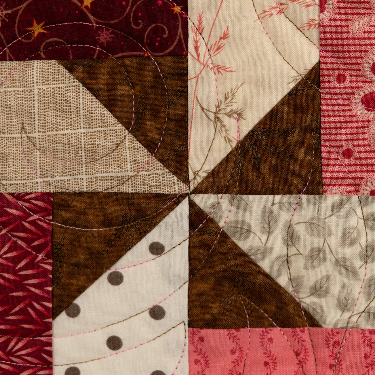 A square quilted fabric block pattern featuring eight distinct patterned sections in shades of red, pink, brown, beige, and white, arranged in a pinwheel-like layout with visible quilting stitches.