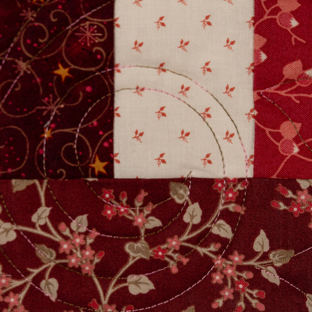 A close-up of a quilted fabric patchwork featuring various patterns in deep red, burgundy, and cream, including floral motifs, stars, and swirls.
