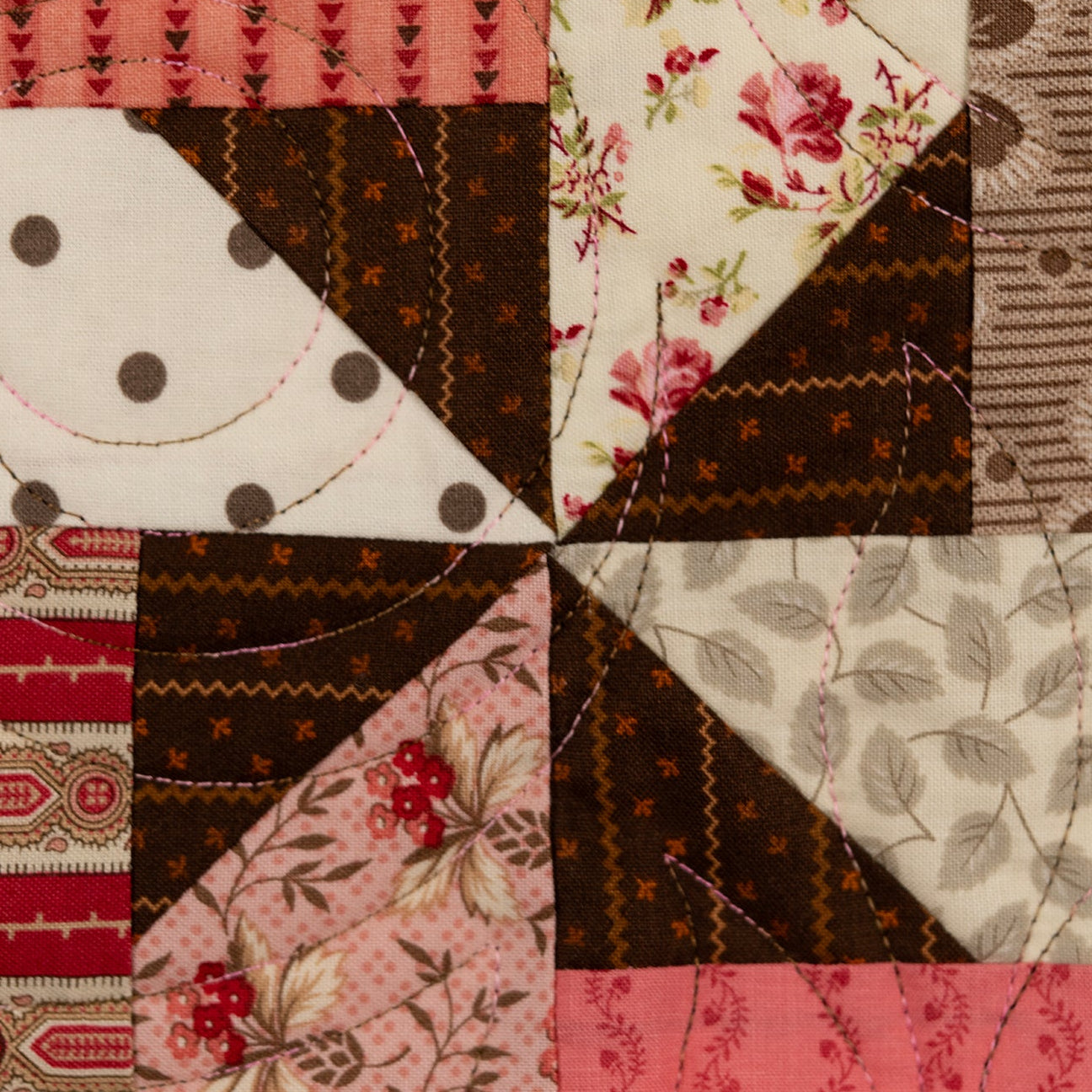 A close-up of a patchwork quilt featuring various fabric patterns including floral prints, polka dots, stripes, and leaf motifs in shades of brown, pink, red, and cream.