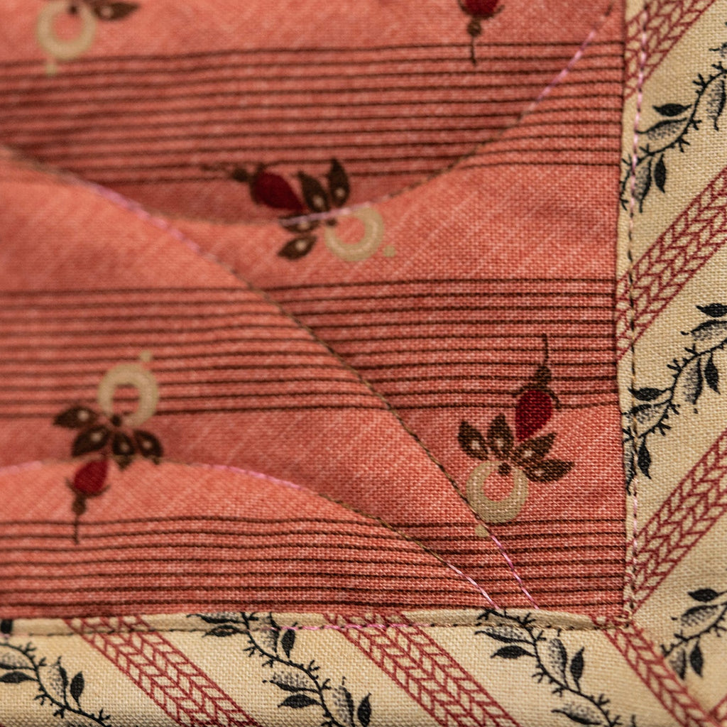 A close-up of a quilted fabric panel in a warm coral pink color, featuring a subtle horizontal stripe pattern and small red and cream floral motifs. The fabric is finished with a decorative border in cream and pink with black leafy details.