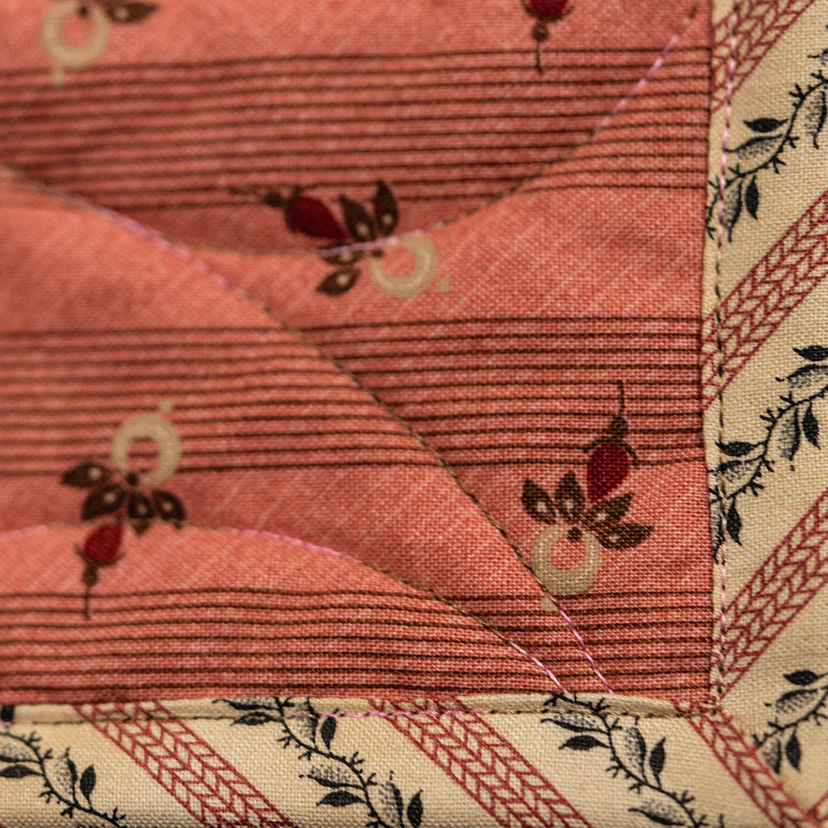 A close-up of a quilted fabric panel in a warm coral pink color, featuring a subtle horizontal stripe pattern and small red and cream floral motifs. The fabric is finished with a decorative border in cream and pink with black leafy details.
