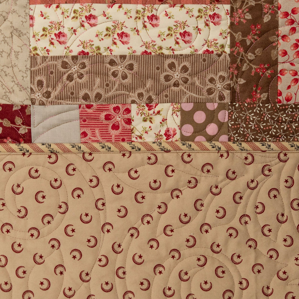 A close-up of a quilted fabric panel featuring a patchwork design with floral and striped patterns in red, pink, brown, and beige, with a large beige section at the bottom showing a repeating red crescent motif.