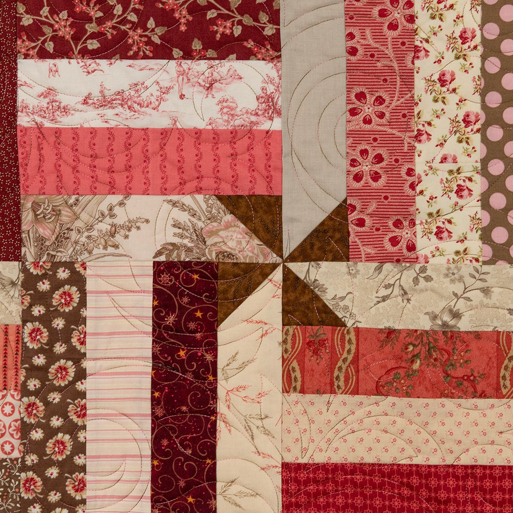 A square quilted textile featuring a patchwork design of red, pink, brown, and cream fabrics with floral, polka dot, striped, and textured patterns arranged in a windmill layout.