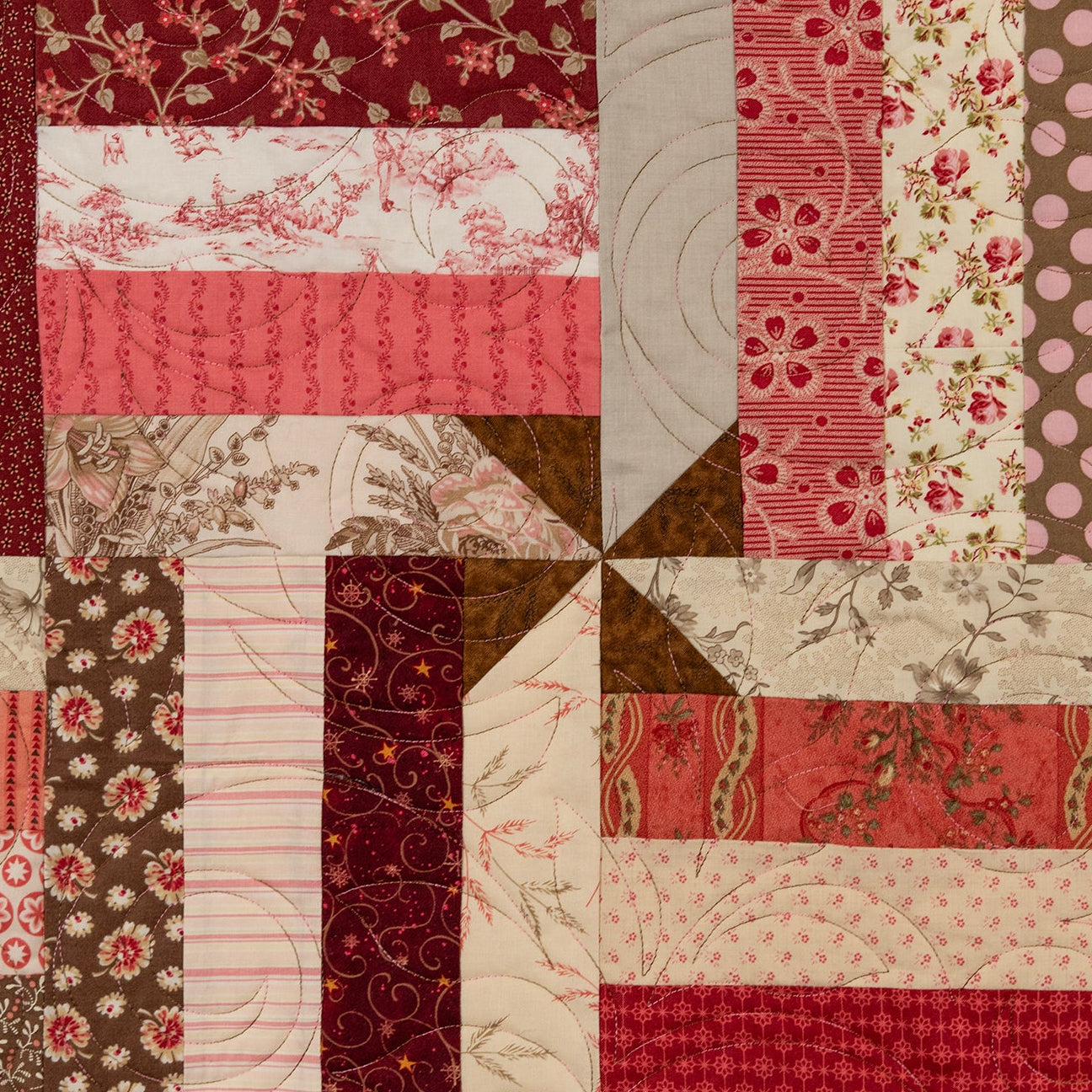 A square quilted textile featuring a patchwork design of red, pink, brown, and cream fabrics with floral, polka dot, striped, and textured patterns arranged in a windmill layout.