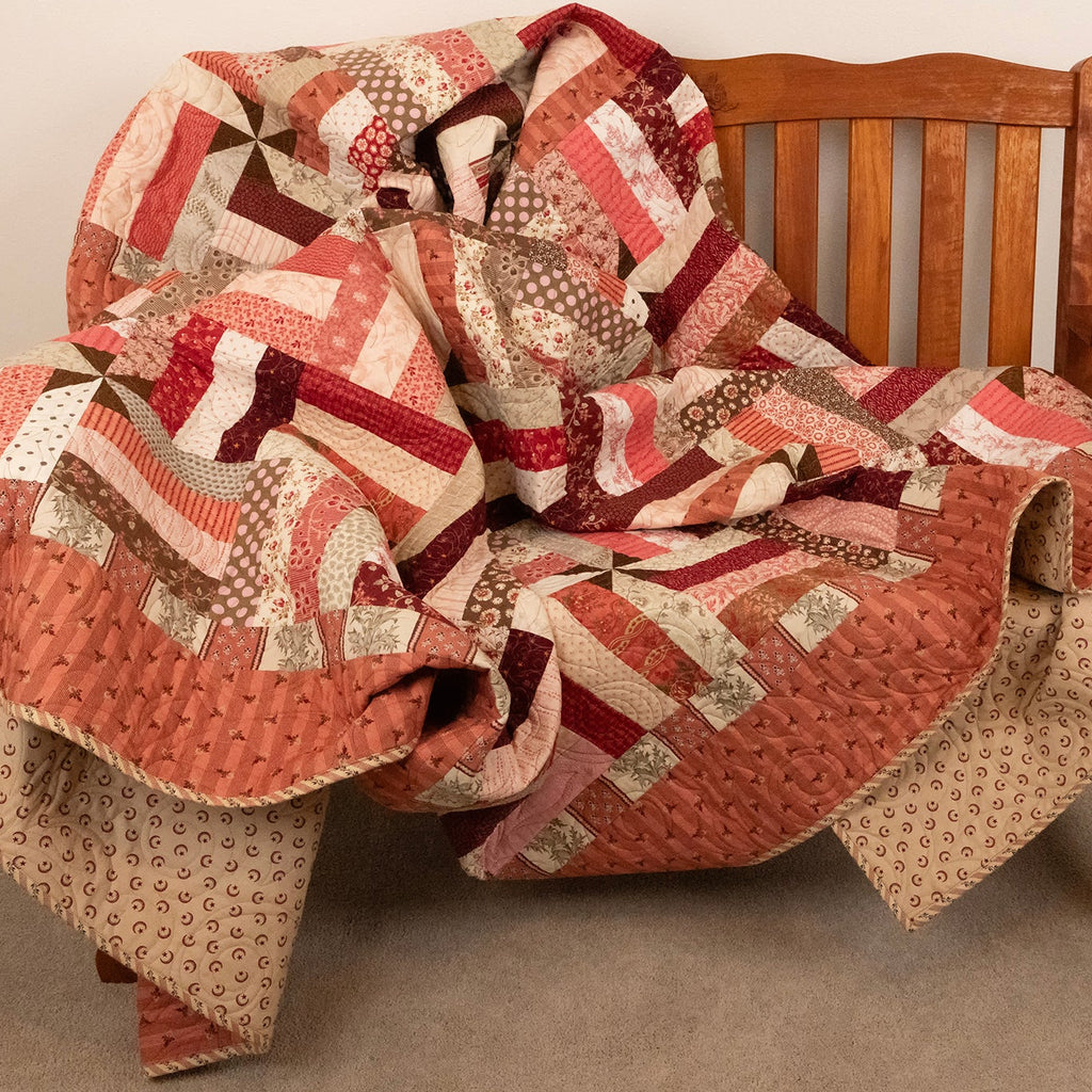 A patchwork quilt in warm red, beige, and brown tones draped over a wooden bench in an indoor setting.